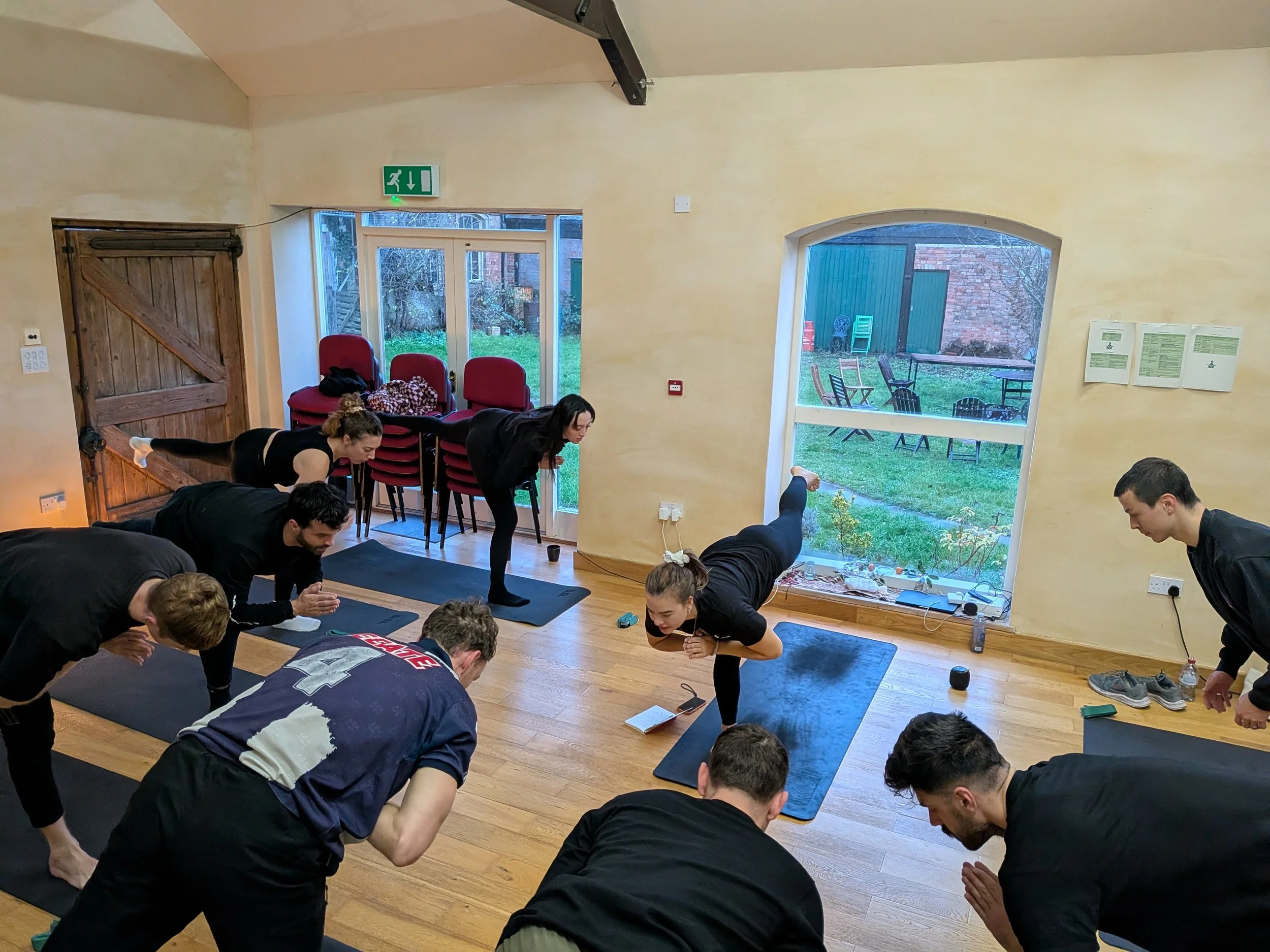 Group of people participating in a yoga or fitness class indoors, performing plank pose on yoga mats, with large windows showing a backyard with chairs and garden.