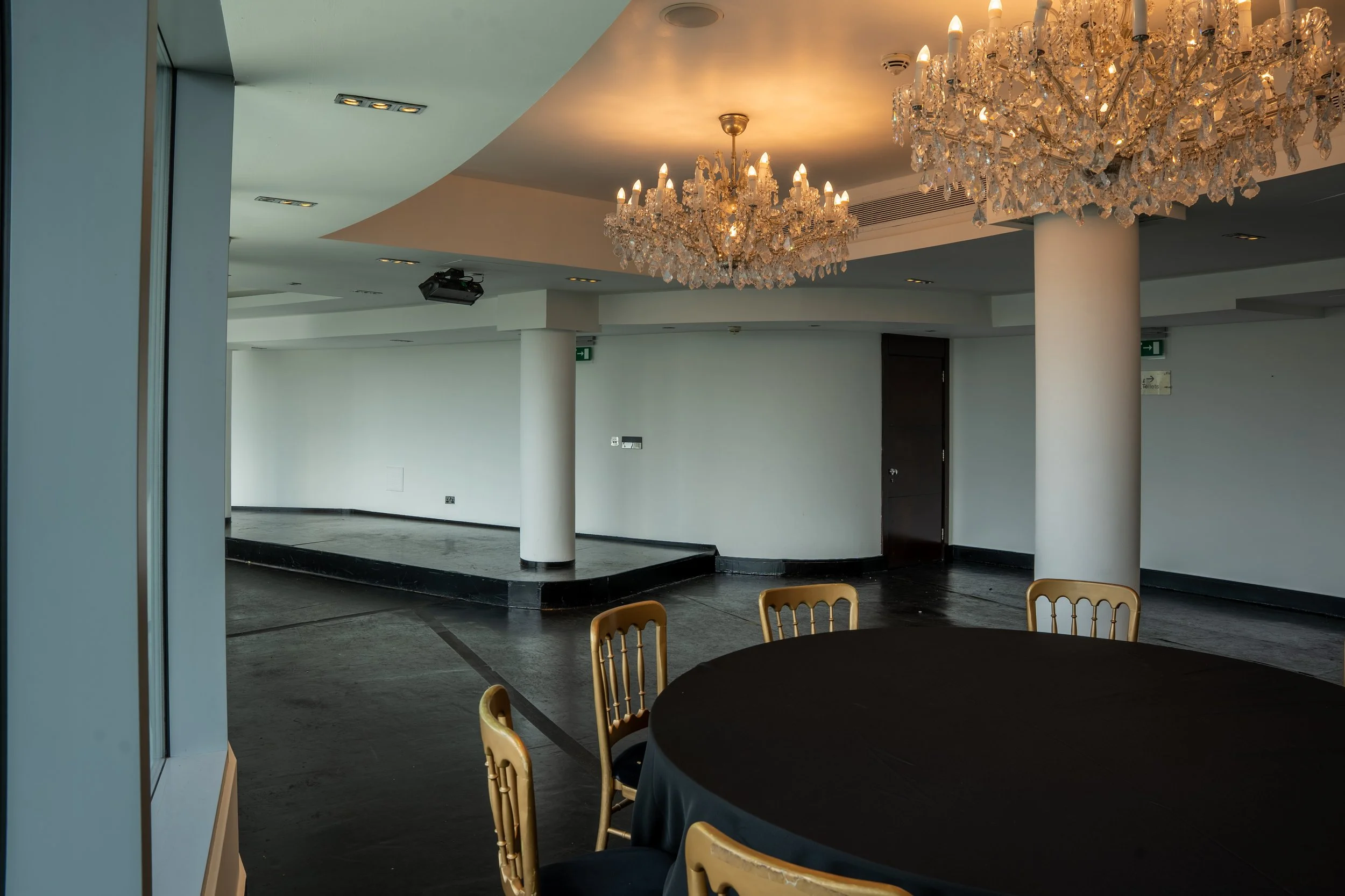 Large, elegant crystal chandeliers hanging from the ceiling in a spacious room with white walls and dark flooring. Part of a round table with gold chairs is visible in the foreground.
