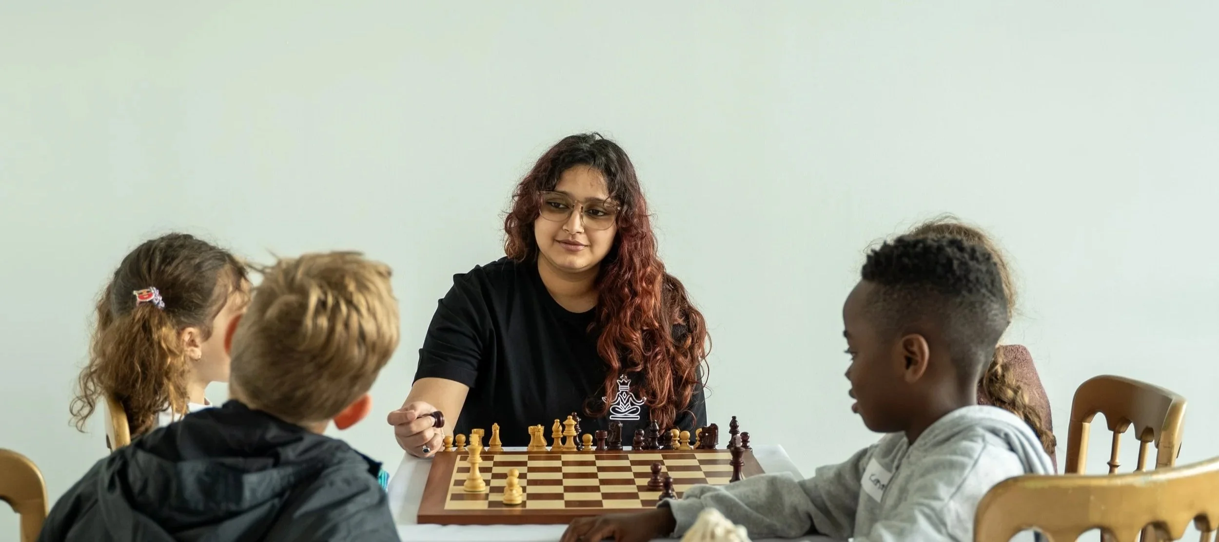 A woman with long, curly red hair and glasses plays chess against four children, seated at a table in a room with a plain white wall.