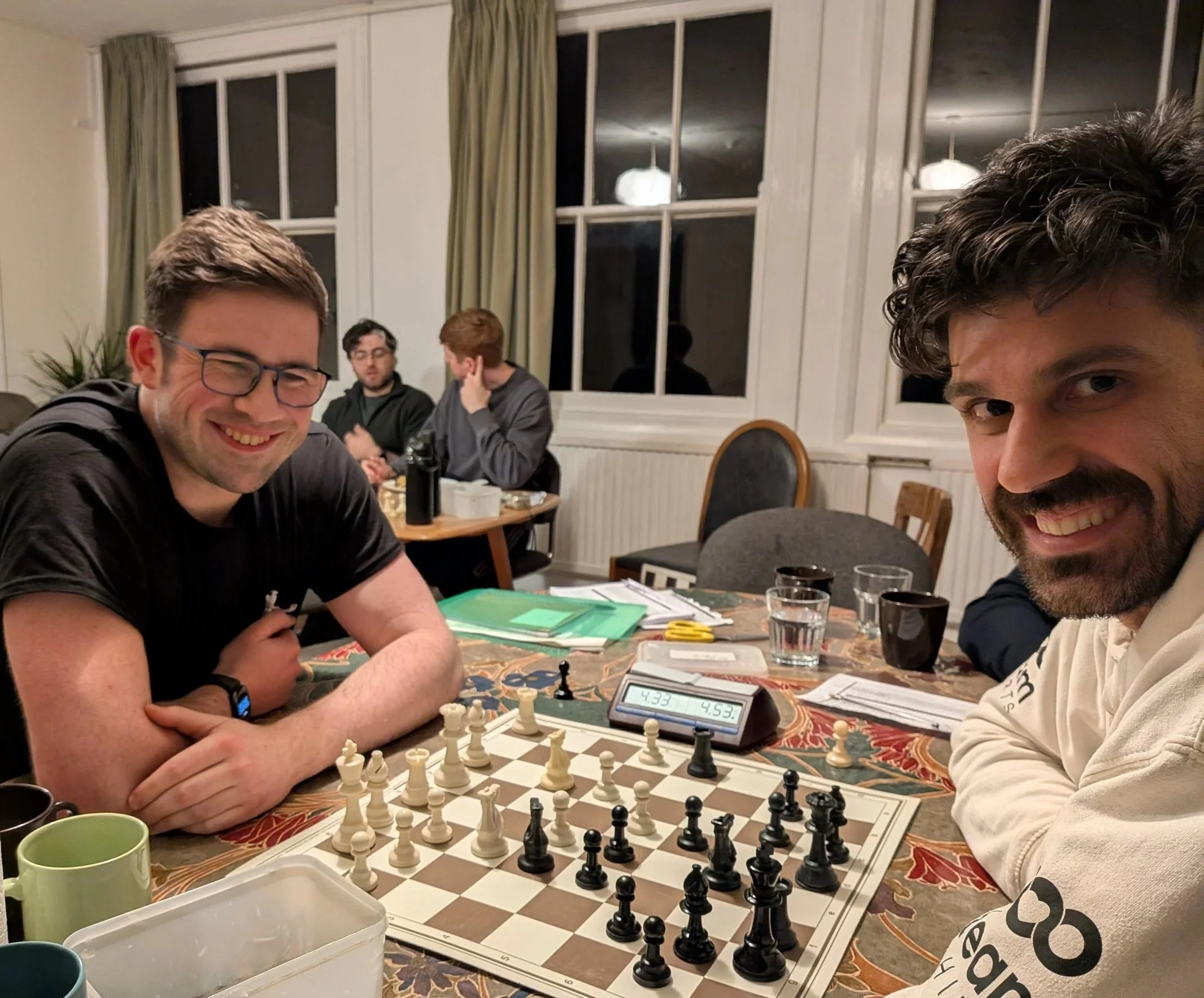 Four young men gathered around a chessboard in a cozy room, playing chess. Two men are sitting at the near side, smiling at the camera, while two others sit in the background, engaged in conversation. The room features large windows with curtains and