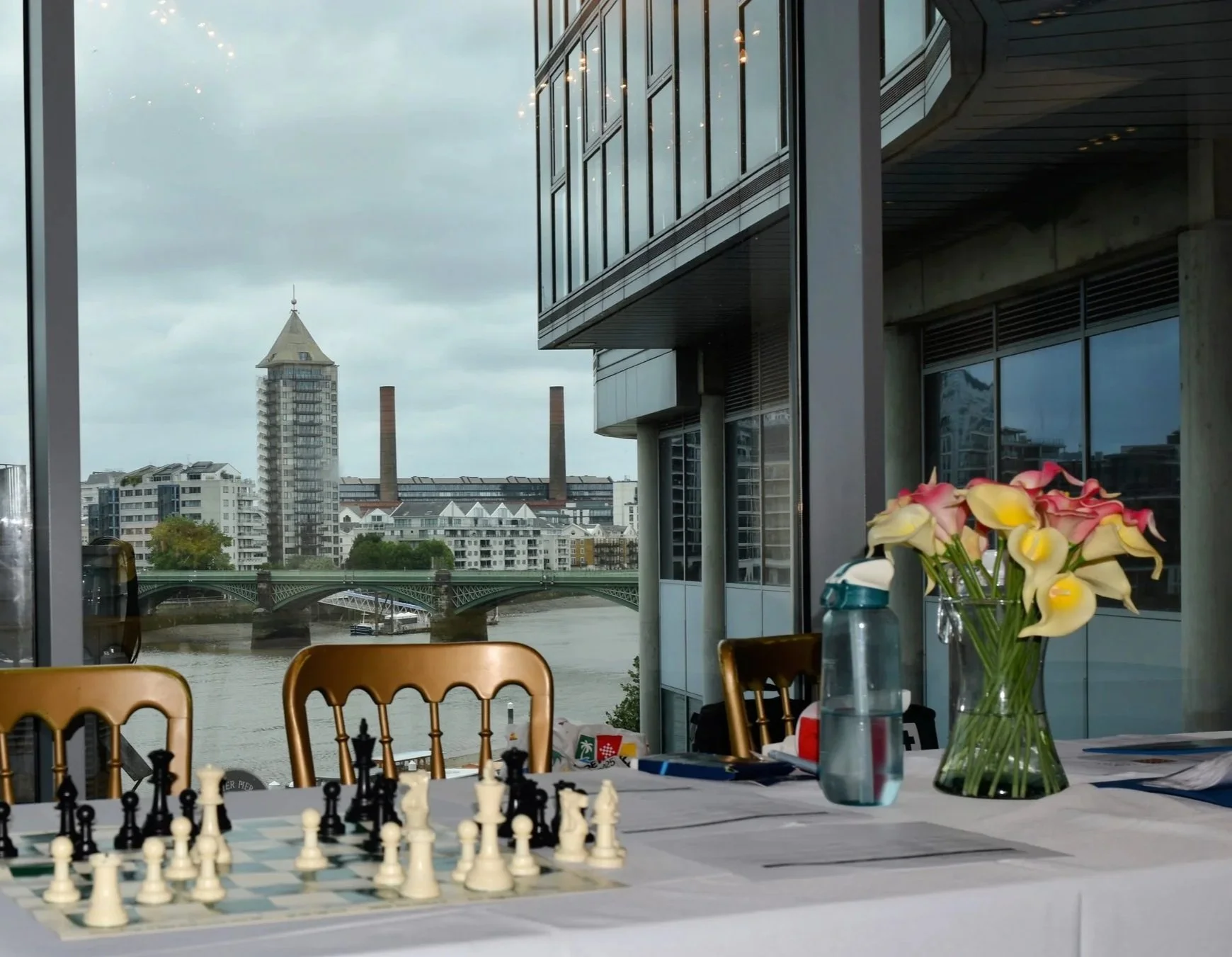 Indoor room with large windows overlooking a cityscape with modern buildings, a river, and a bridge; a table set up with a chessboard and pieces; a bouquet of pink and white calla lilies in a glass vase; chairs around the table; cloudy sky outside.