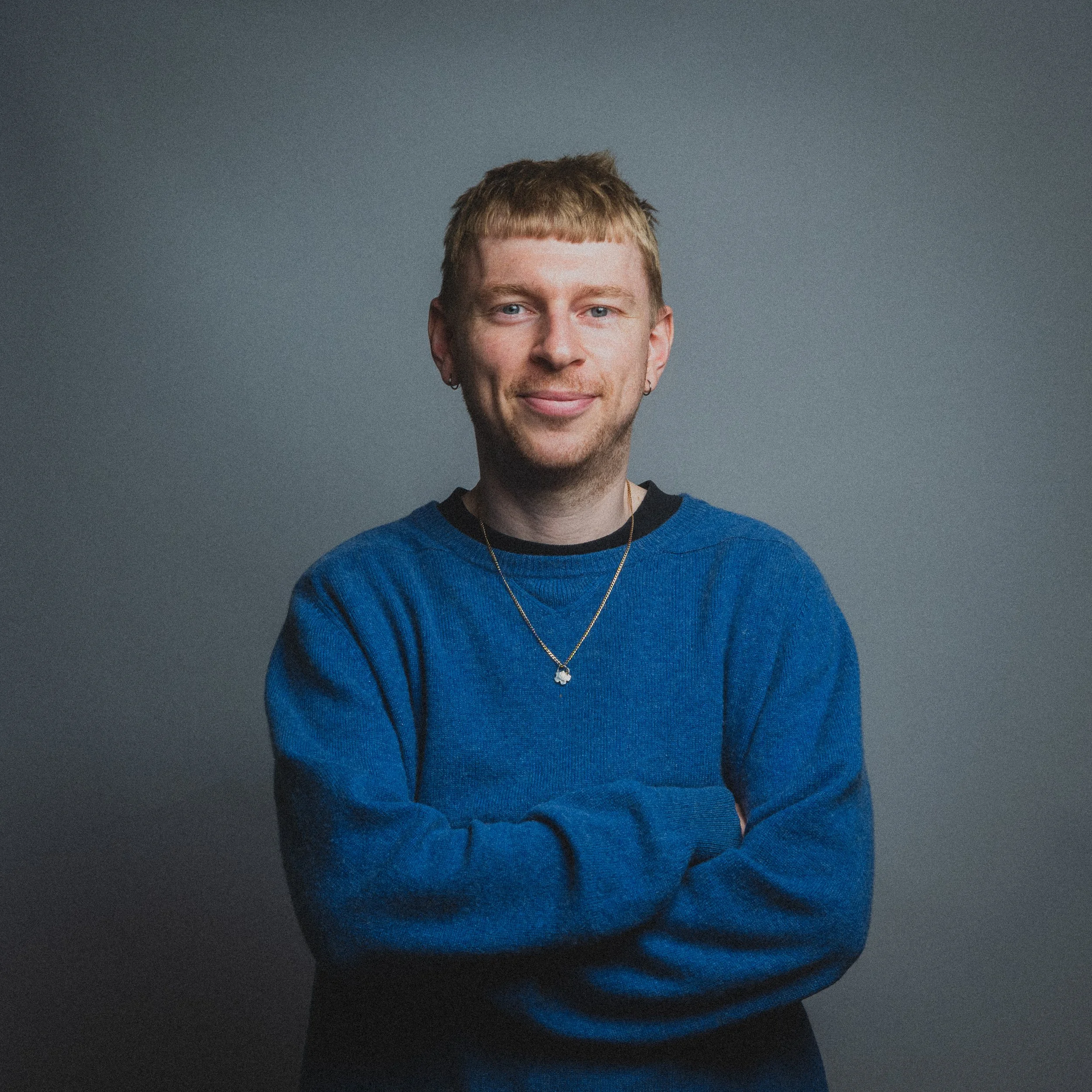 Close-up of a young man with blond hair, blue eyes, and a small earring, wearing a blue sweater and a necklace, against a gray background.