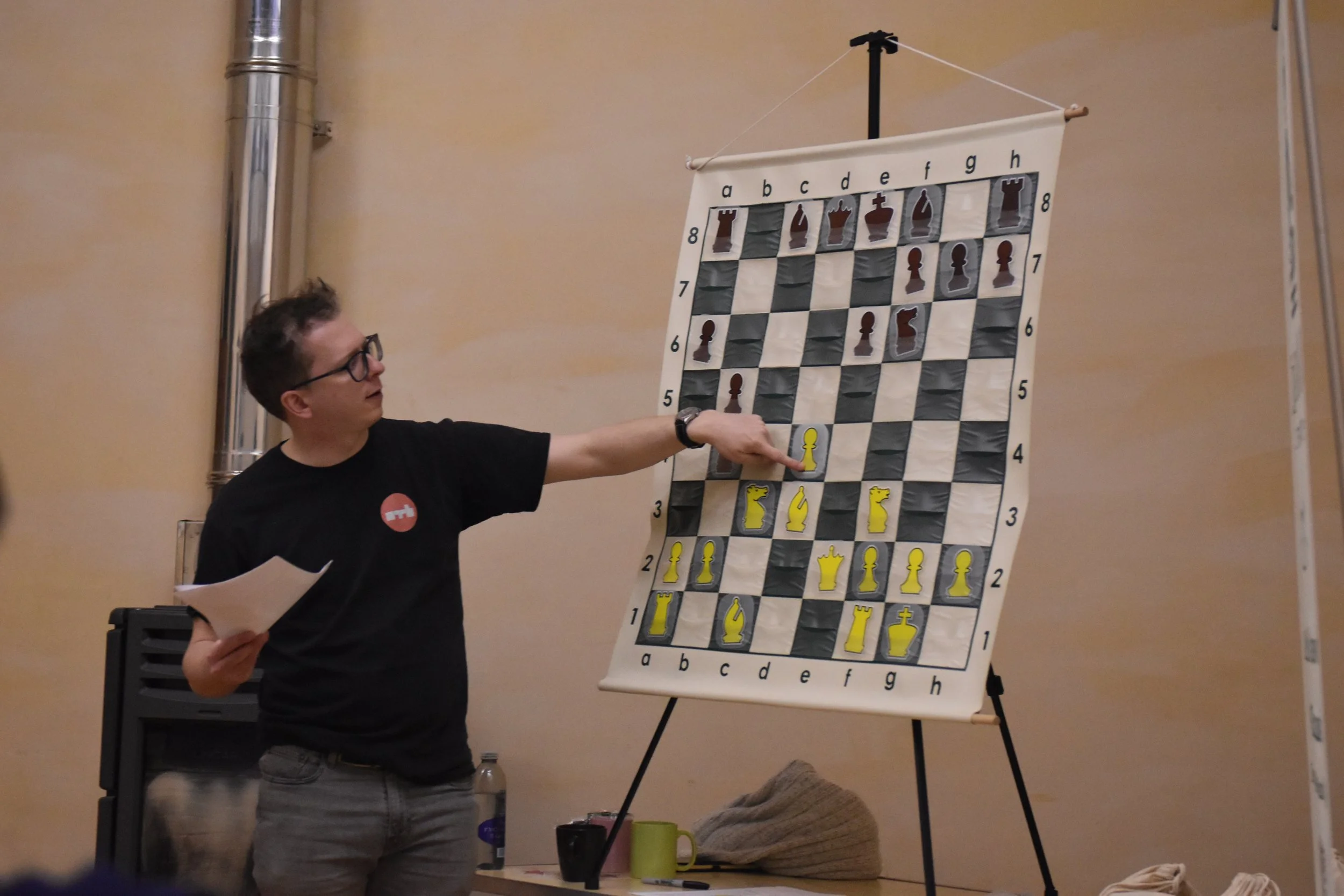 A man with glasses and a black t-shirt pointing at a large chess-themed target on a stand, with chess pieces illustrations and coordinates, in a room with beige walls and a metal pipe.