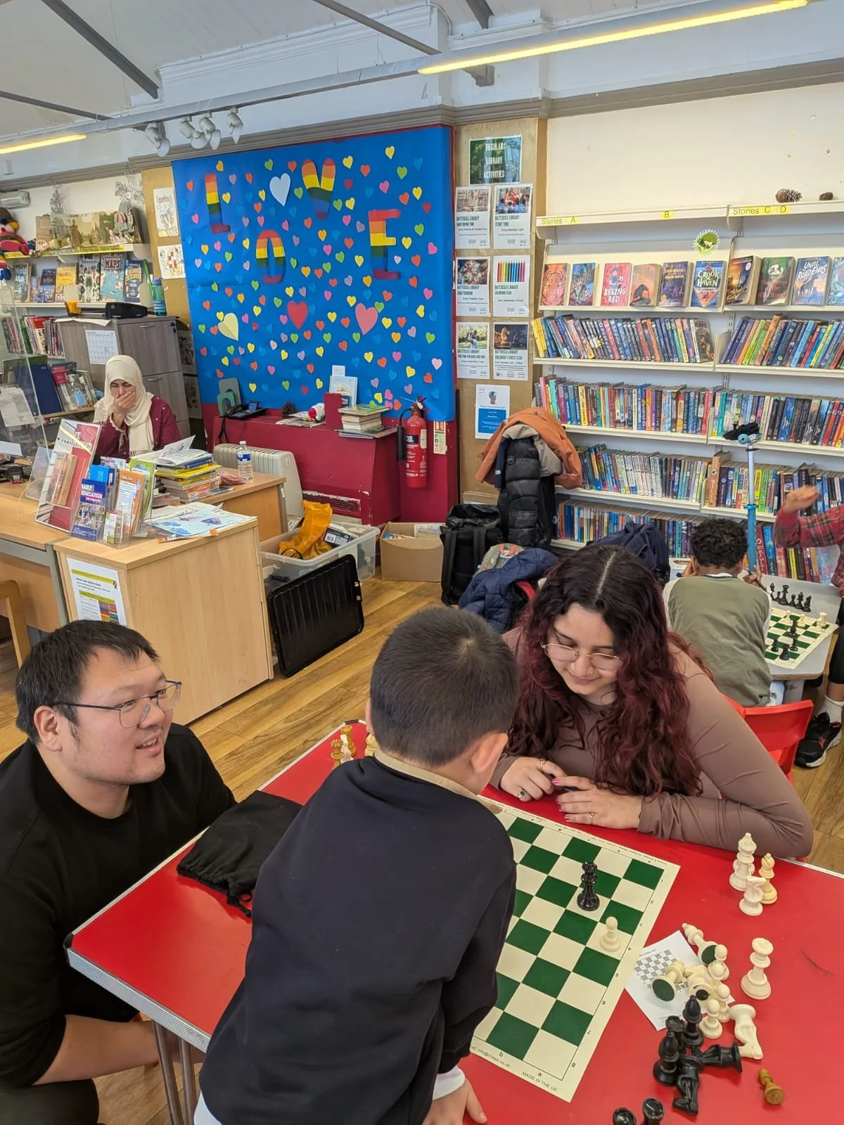 People playing chess at a bookstore or library with shelves of books and a colorful heart-themed decoration on the wall reading 'LOVE'.