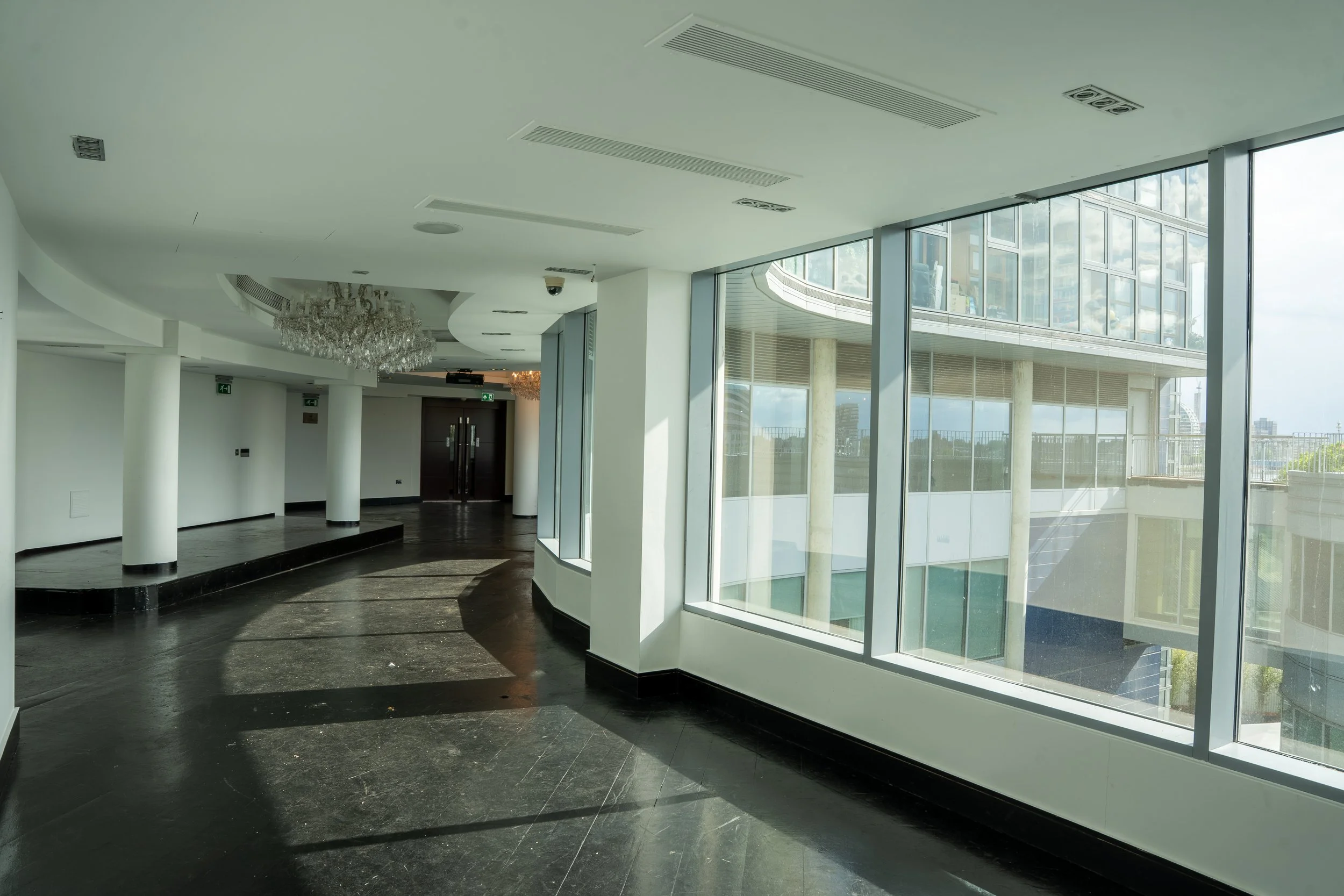 Modern interior space with large floor-to-ceiling windows, reflecting city buildings, with black marble flooring, white walls, and decorative chandeliers.