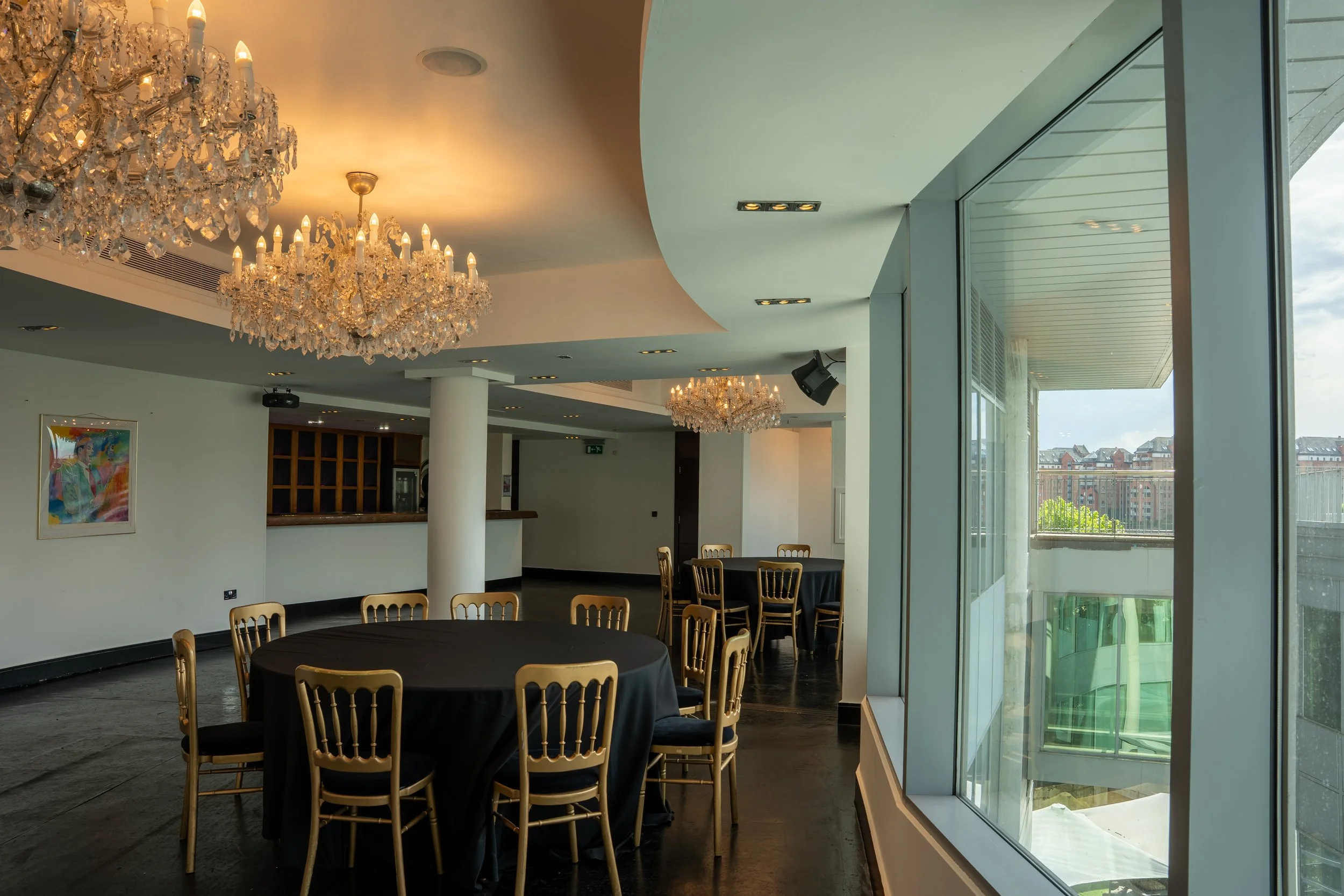 Interior of a modern banquet hall or event space with large crystal chandeliers hanging from the ceiling, round tables with black tablecloths, and gold-colored chairs. There is a large window on the right side showing a view of buildings and trees outside, and some artwork on the wall.