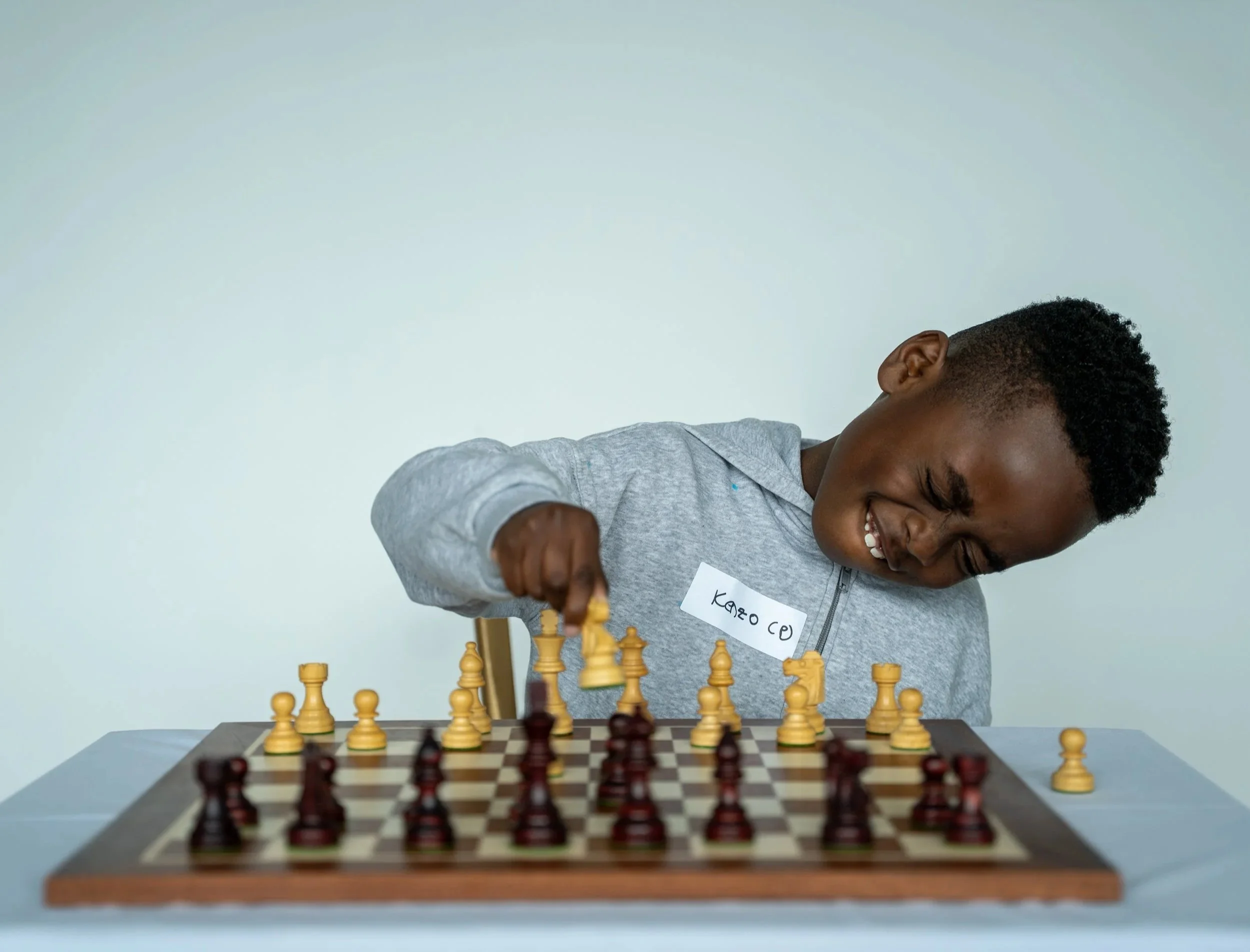 Young boy playing chess at a table, smiling, wearing a gray hoodie, with a white name tag on his chest and short curly hair.