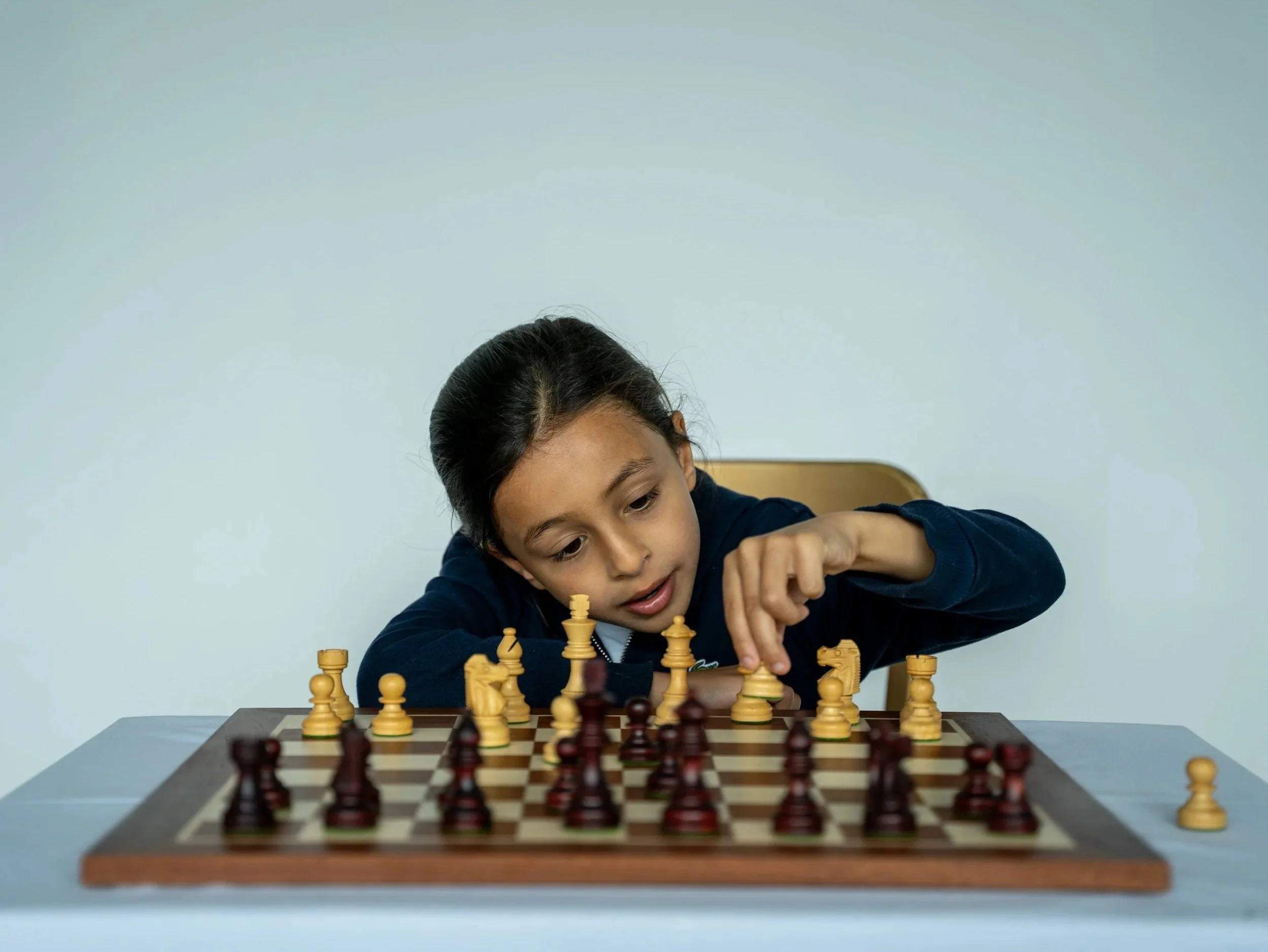 Young boy playing chess at a table, focused on the game, with a light blue wall in the background.