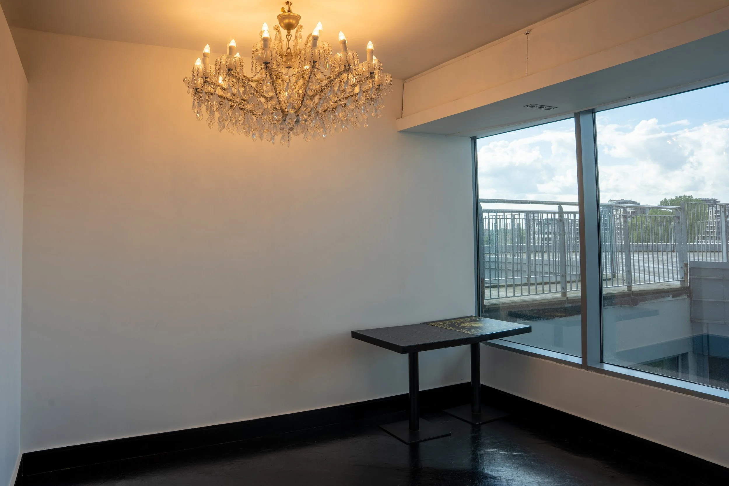 A modern, minimalist room with a large crystal chandelier hanging from the ceiling, a sliding glass door leading to a balcony, and a small black table with a patterned mat on top.