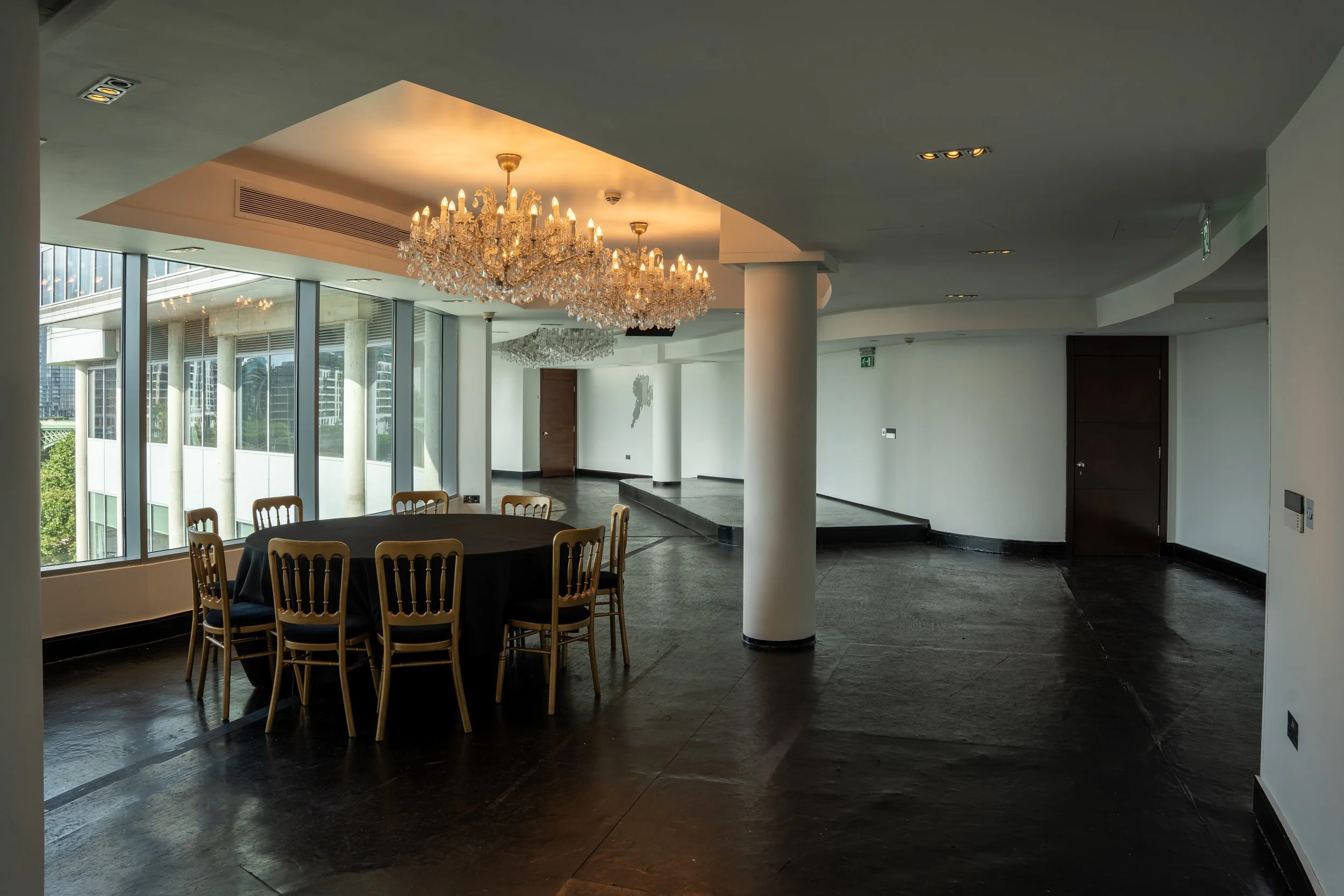 Interior of a modern banquet or conference room with large windows, crystal chandeliers, a round table with a black tablecloth and gold chairs, and white walls with minimal decor.