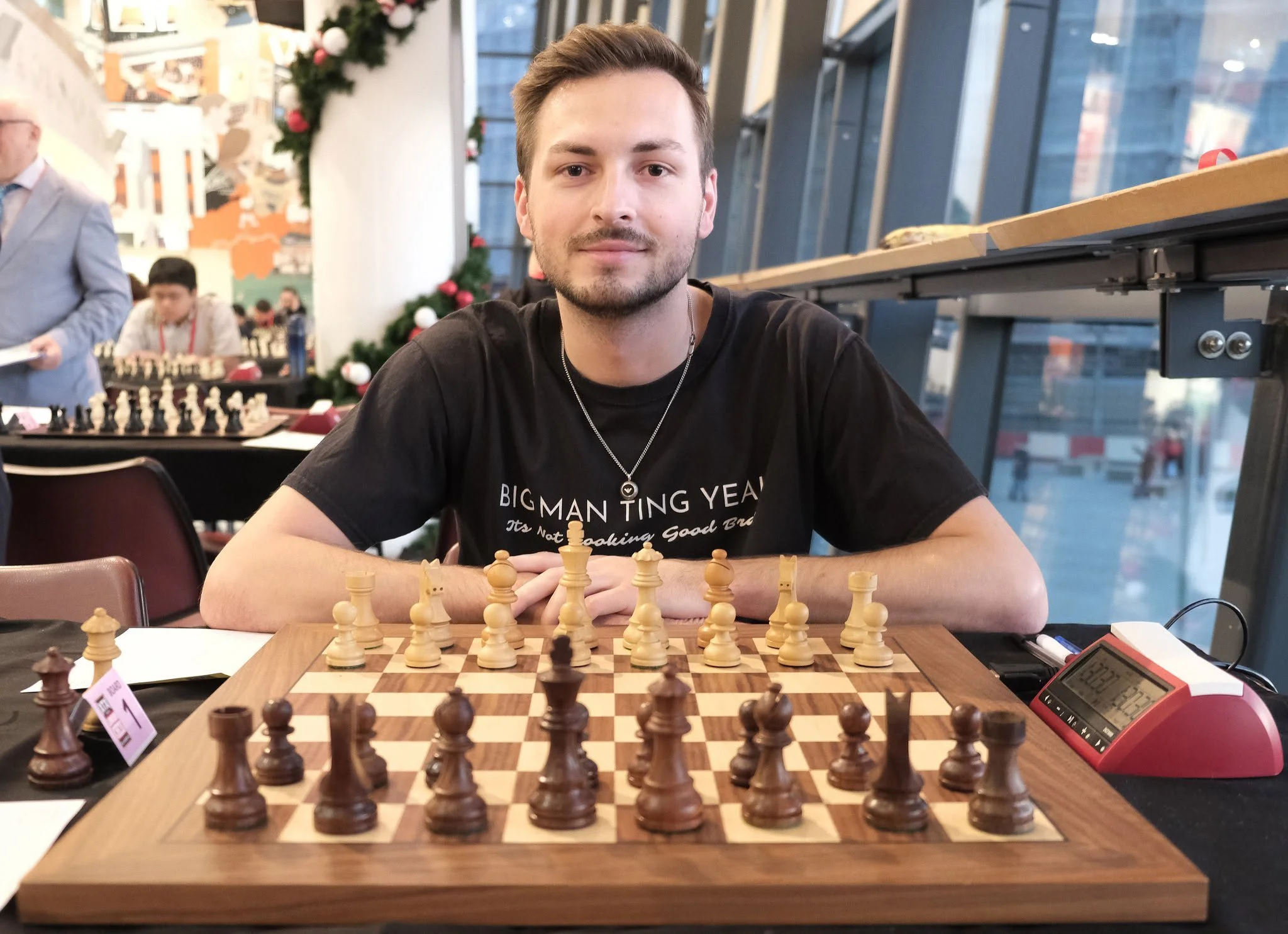 A young man with light brown hair and a beard, wearing a black T-shirt, sits at a chessboard looking at the camera inside a modern indoor space with large windows and decorations. There are other people in the background, some playing chess, some standing, and Christmas decorations are visible.