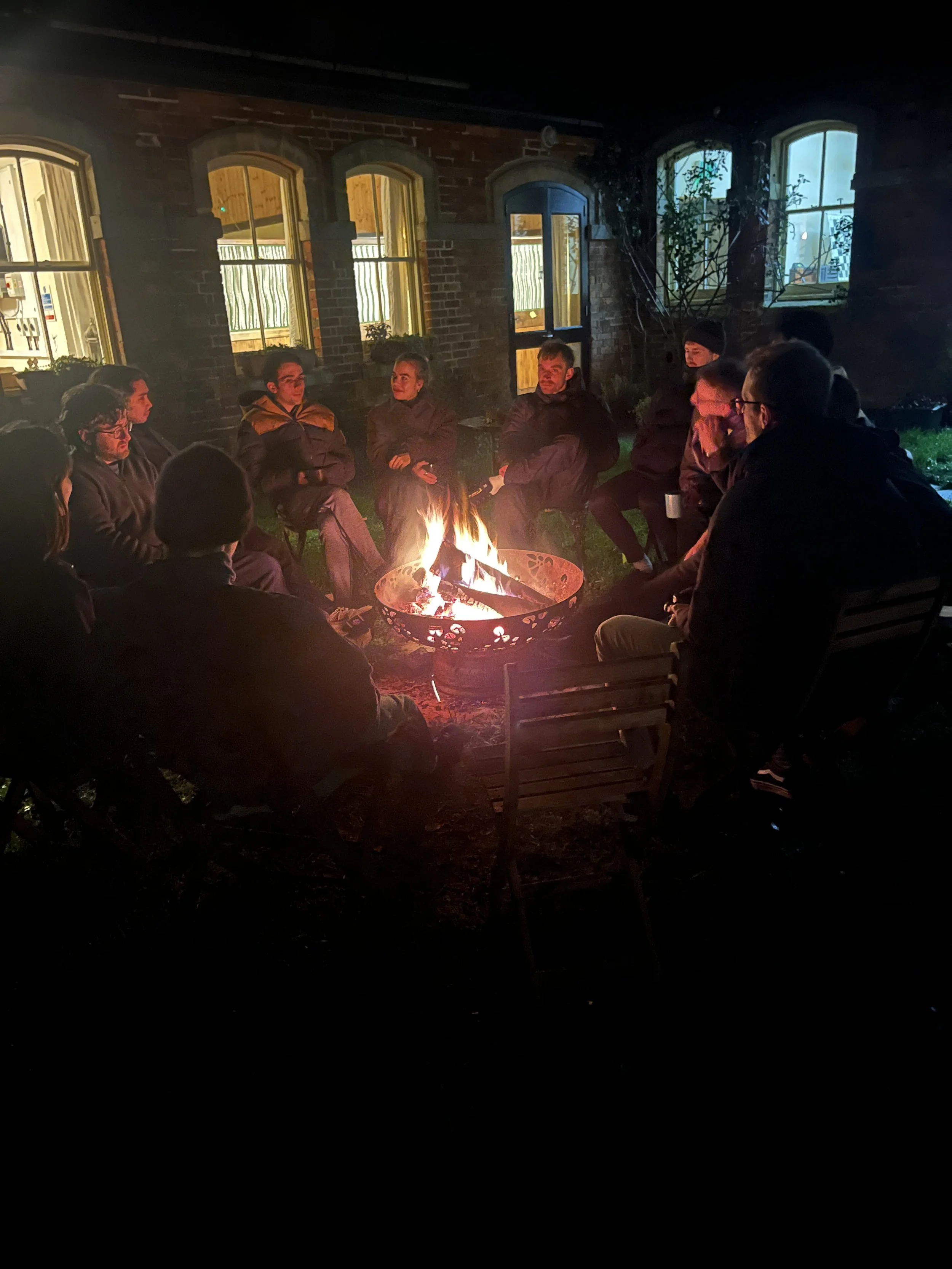 People sitting around a campfire outside at night, near a brick building with illuminated windows and a door.