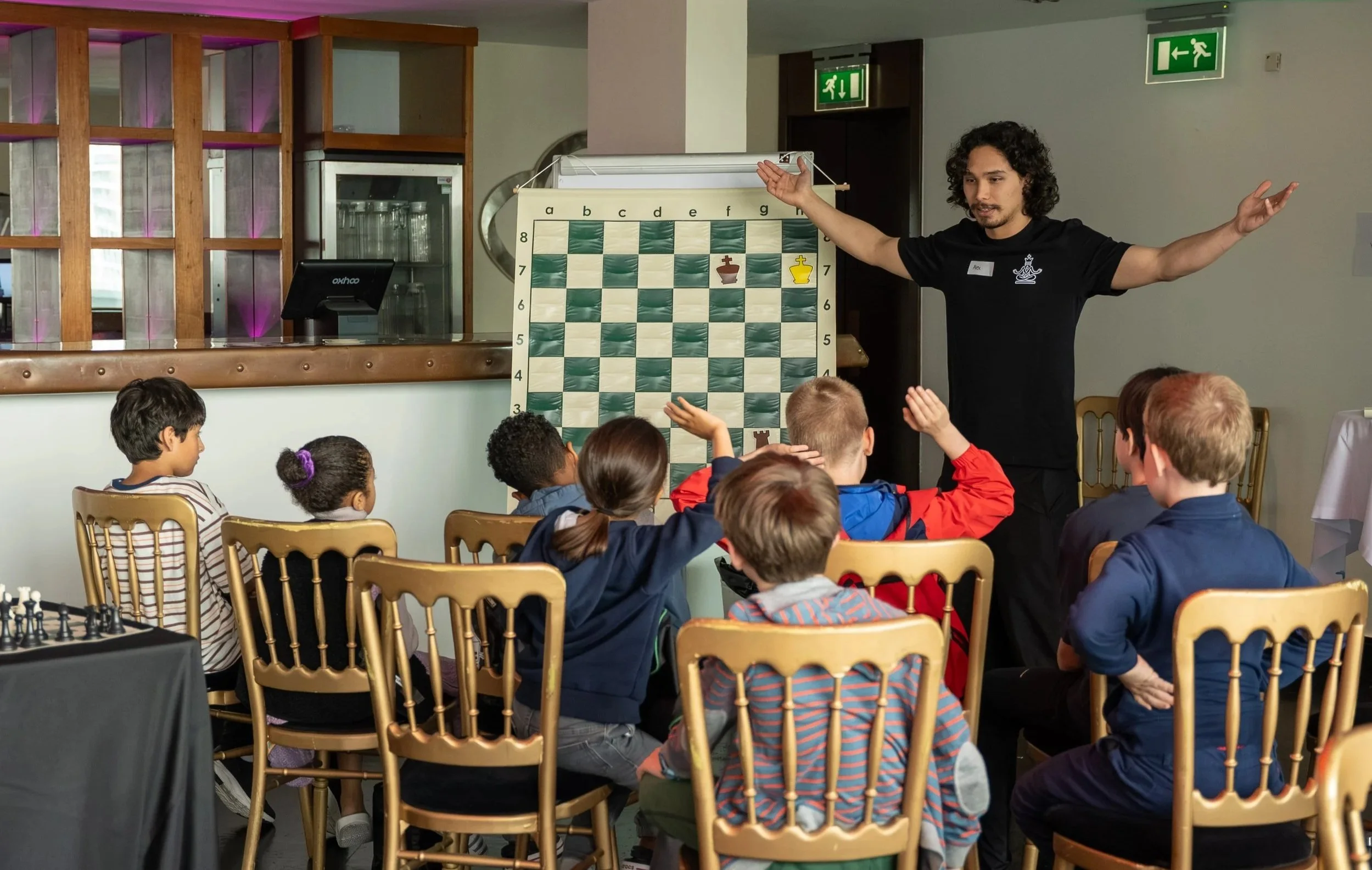 A man giving a chess lesson to children in a classroom or cafe, with a large chessboard and pieces on a stand in front of him.