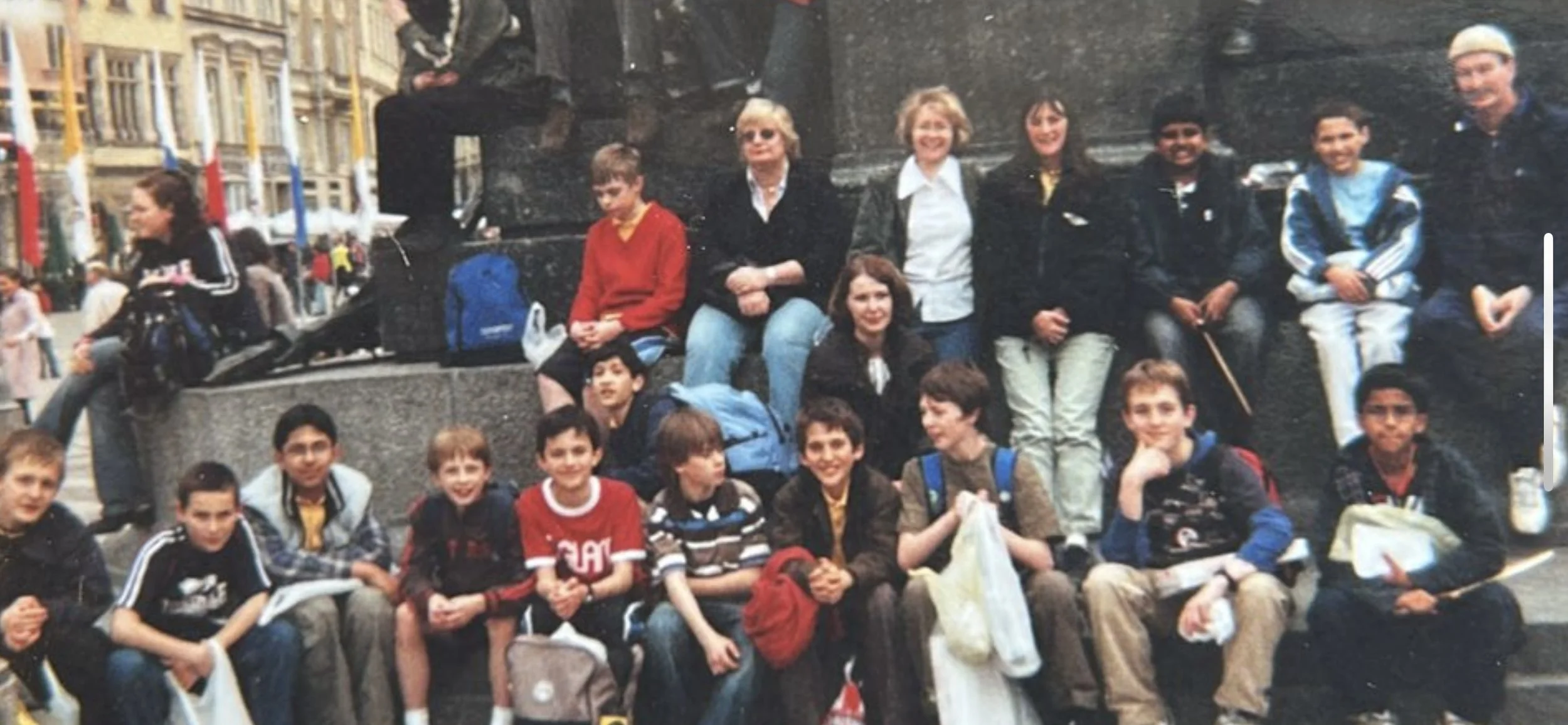 A group of children and a few adults sitting and standing on a city square in front of a black monument. The children have backpacks and are smiling or looking at the camera. The background shows buildings and a busy street.