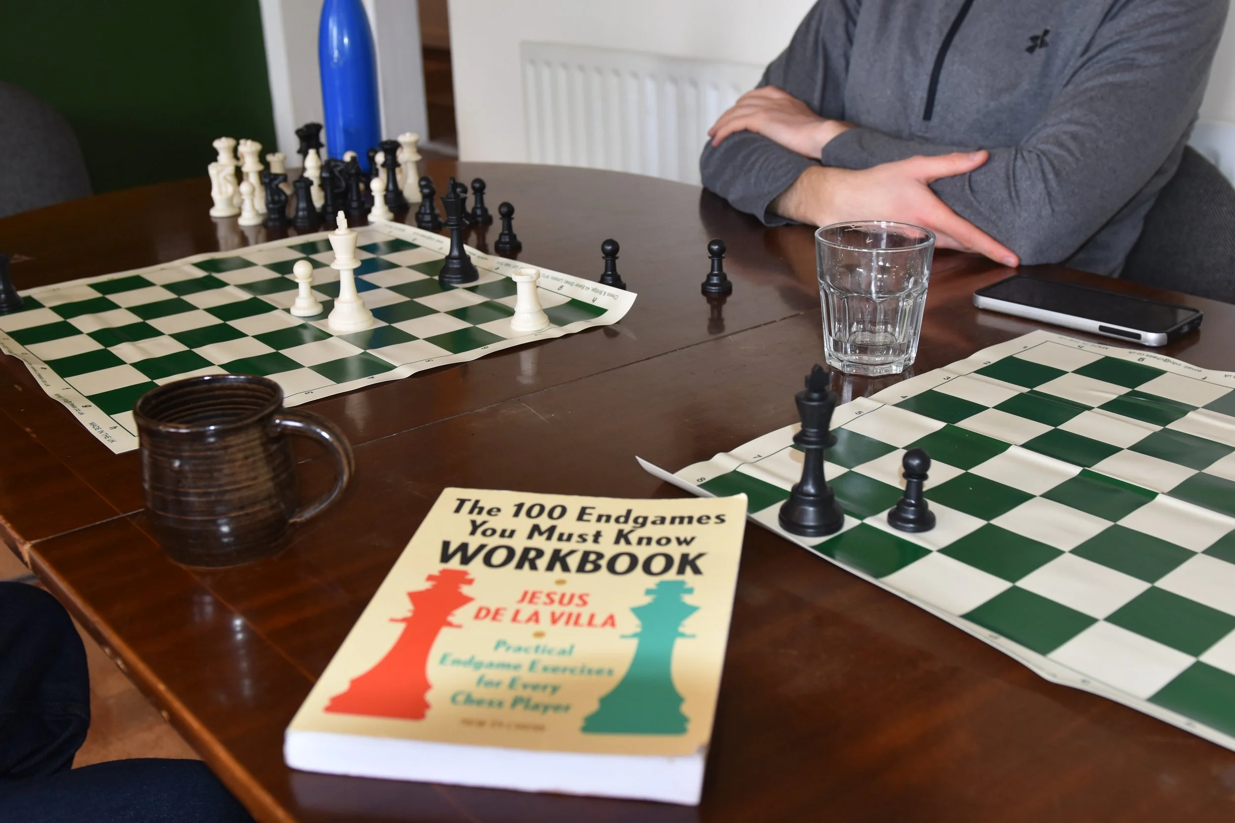 A chess game set up on a wooden table with a glass of water, a smartphone, and a booklet titled 'The 100 Endgames You Must Know Workbook' visible in the foreground.