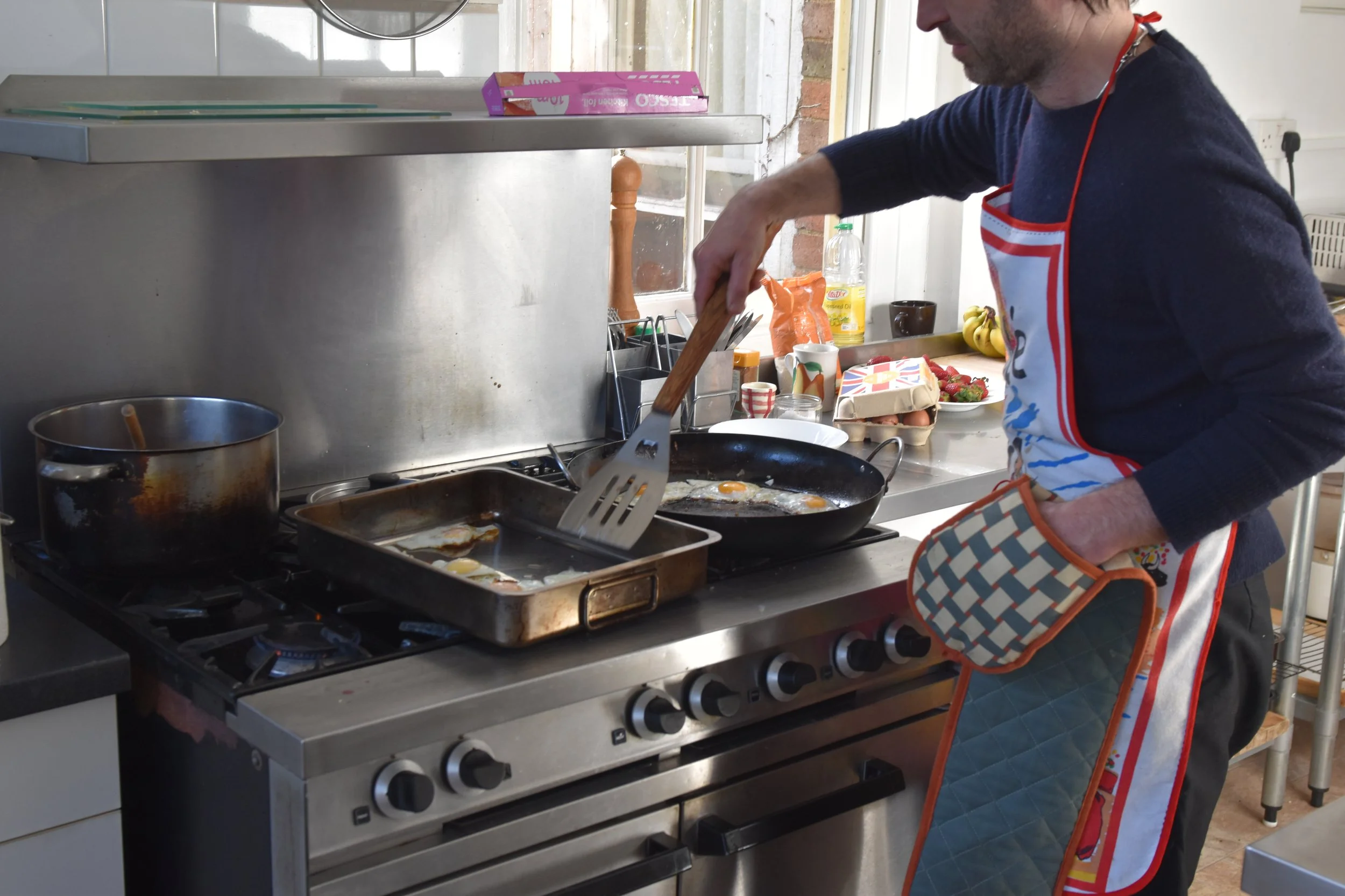 A person cooking breakfast eggs in a frying pan on a kitchen stove, wearing a colorful apron, with kitchen utensils and food items on the countertop nearby.