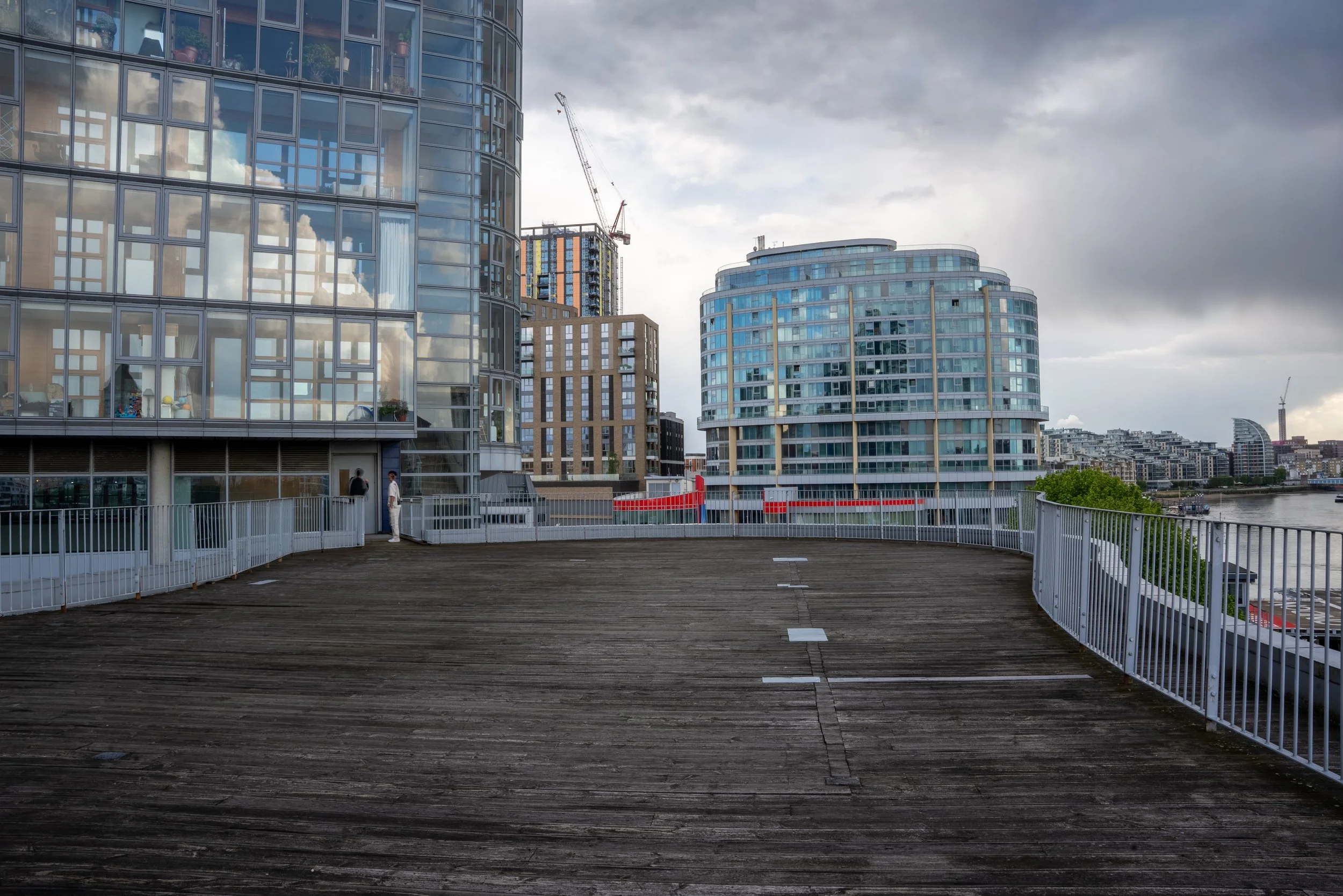 View from a wooden outdoor deck area with white railings, overlooking modern glass buildings and the city skyline under cloudy skies.