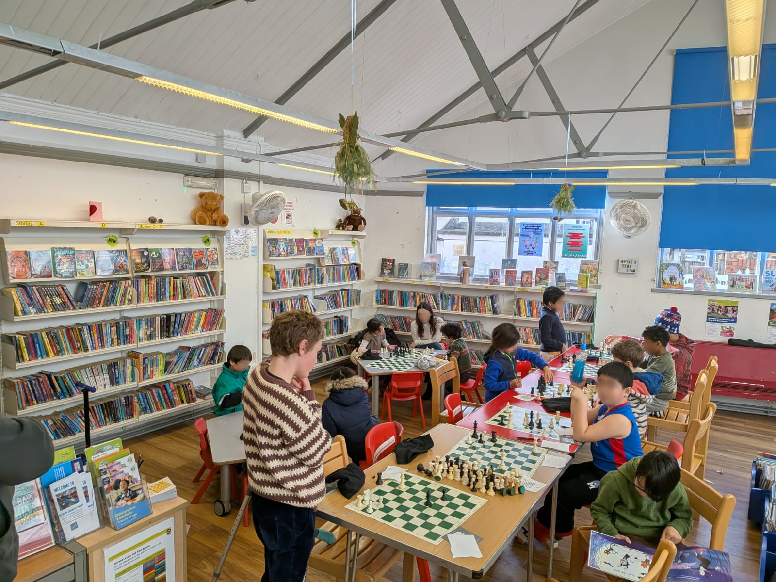 Children and adults playing chess in a library bookstore with shelves of books, posters, and hanging decorations.