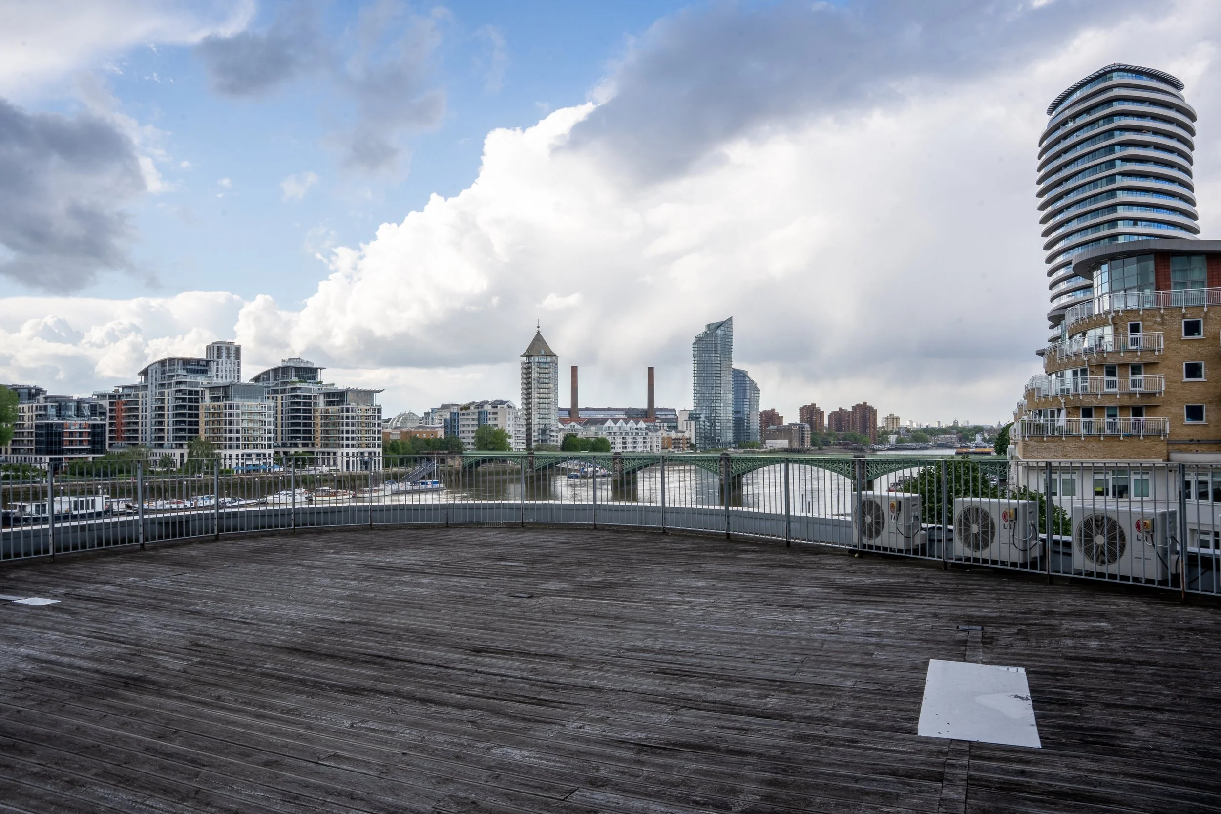 City skyline view of modern high-rise buildings along a waterfront, with wooden deck and railing in the foreground, cloudy sky overhead.