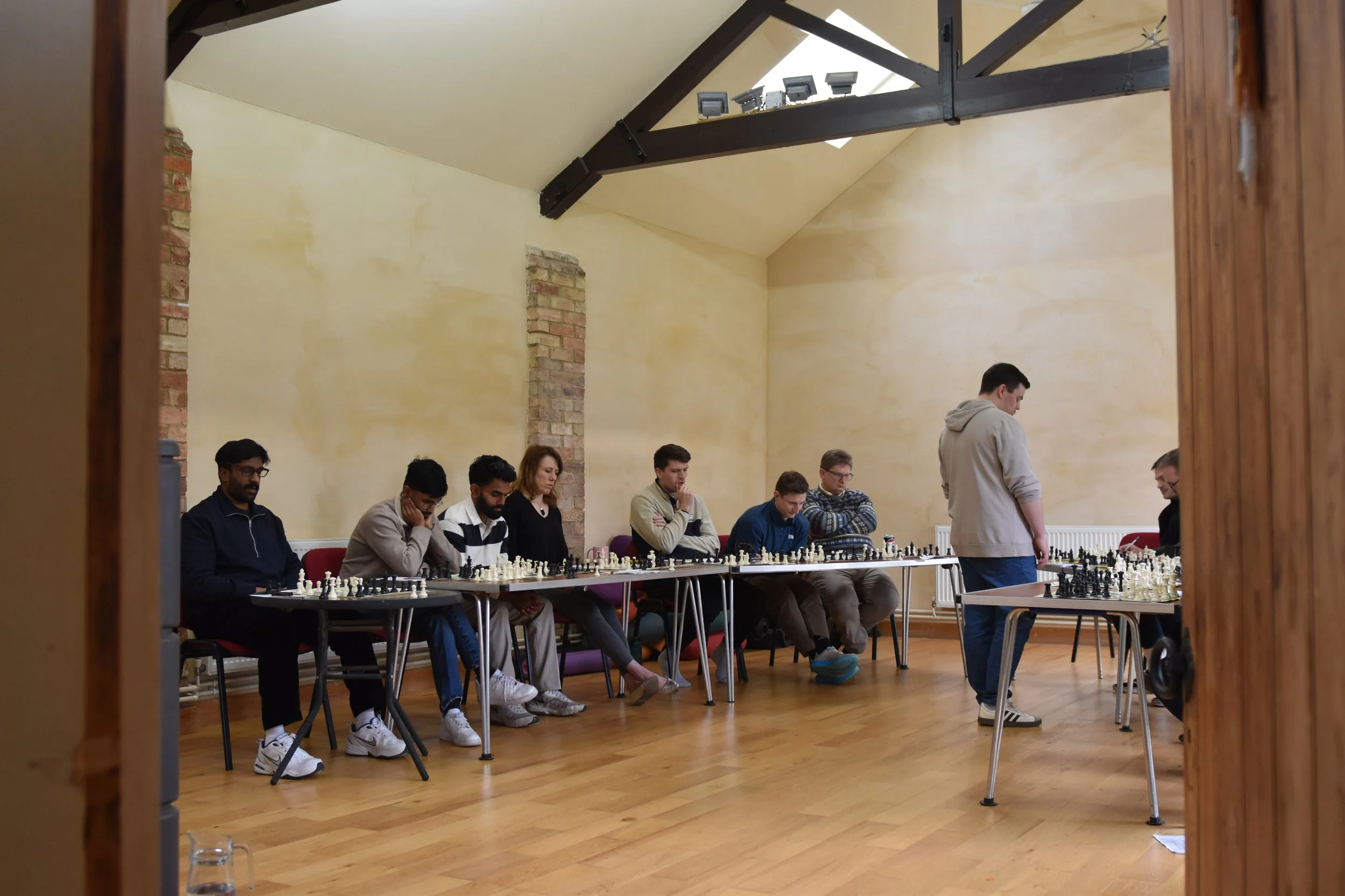 A group of people sitting at tables playing chess in a room with wooden floors and beige walls. Some appear to be concentrating, others are looking at the chessboard. A person in a gray hoodie is standing near a table, possibly observing or officiati