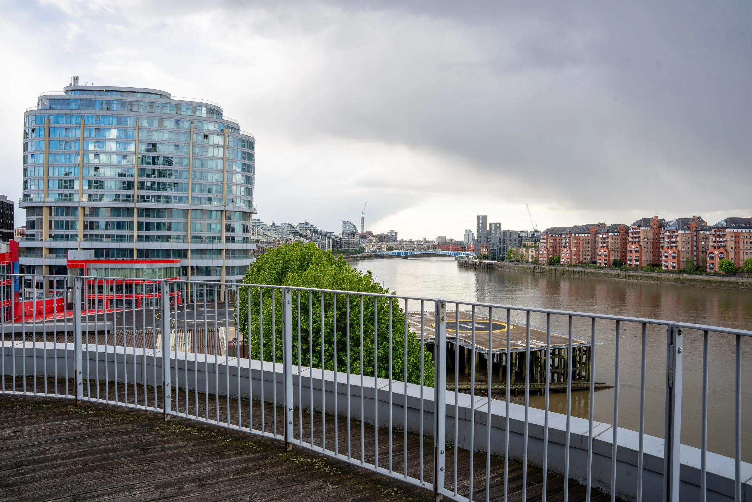 A cityscape view featuring a modern glass building on the left, a river running through the center, and residential buildings on the right, with a cloudy sky overhead.