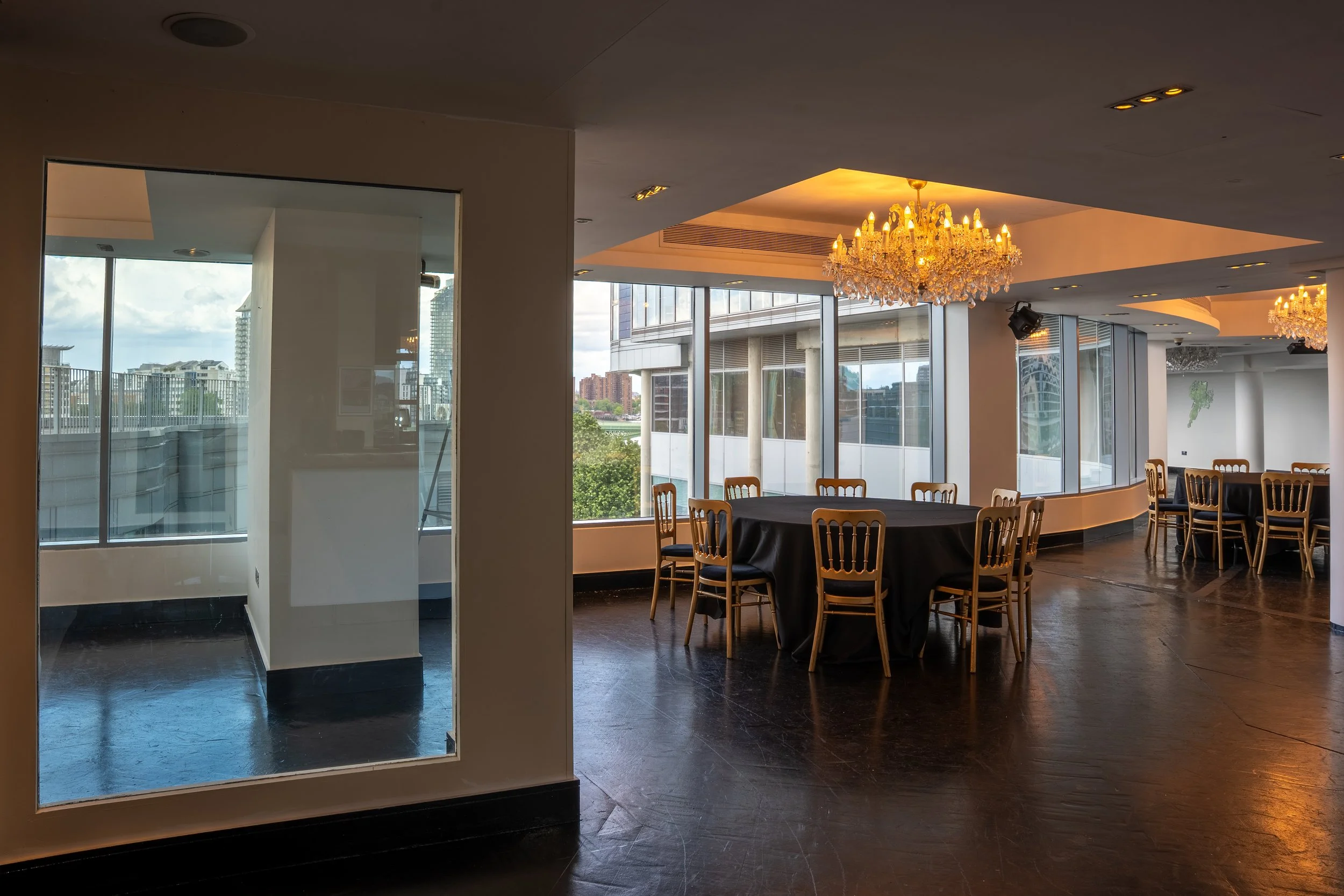 An empty dining area with round tables covered with black tablecloths and wooden chairs, large windows showing city buildings and a cloudy sky, chandeliers hanging from the ceiling, and a glass door leading outside.