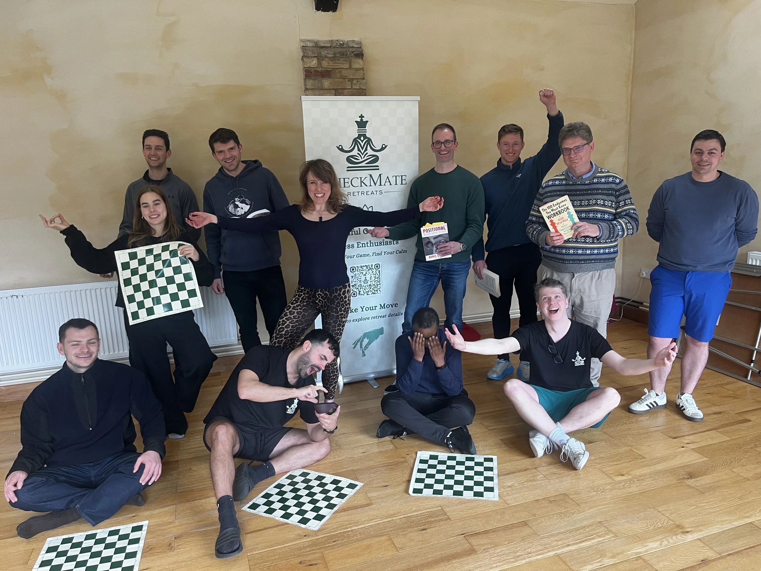 A group of ten smiling people in a room with beige walls, some standing and some sitting on the floor. Several are holding books or chessboards. Behind them is a banner with a chess logo and text, and a small child is finally visible at the bottom of