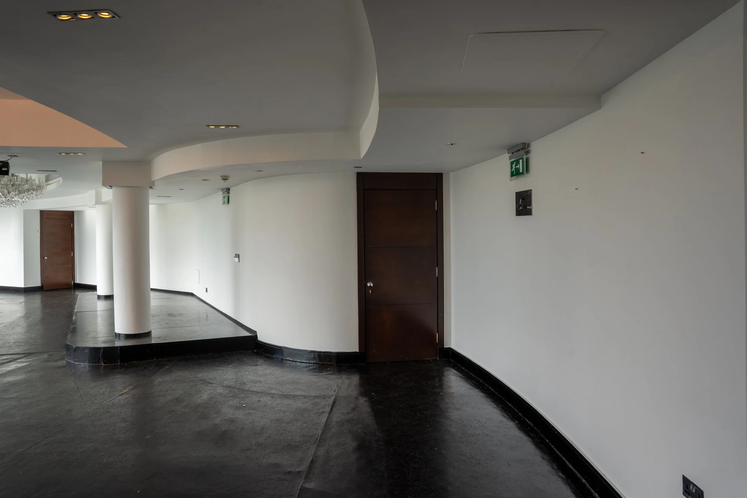 Interior of a modern building corridor with white walls, a dark polished floor, wooden doors, ceiling lights, a green emergency exit sign, and a chandelier partly visible on the left.