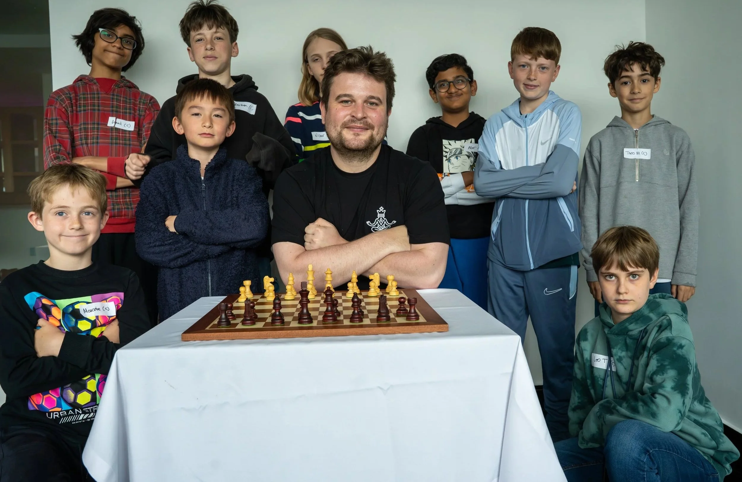 Group of children and a man gathered around a chessboard, with chess pieces set up on it, in a room with plain walls.