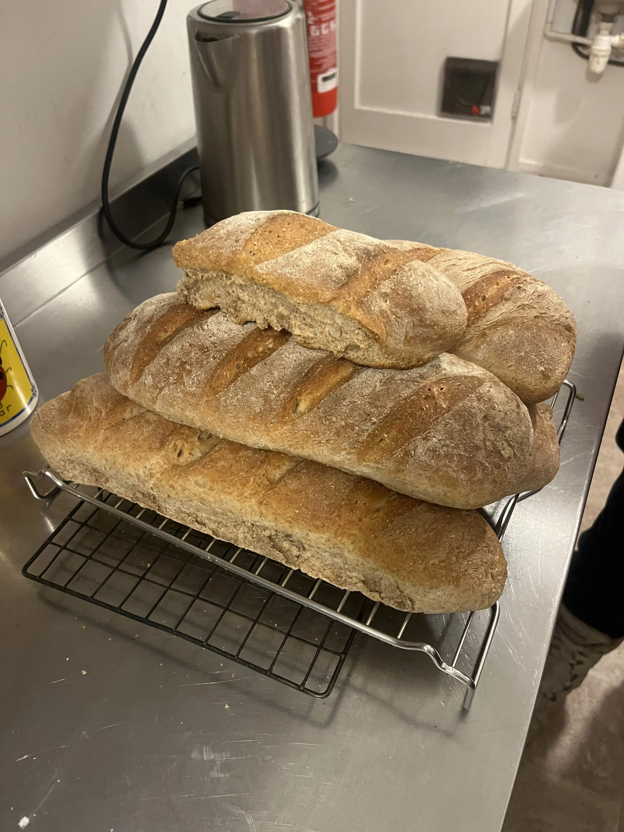 Three loaves of bread on a wire rack on a stainless steel countertop in a bakery or kitchen setting.