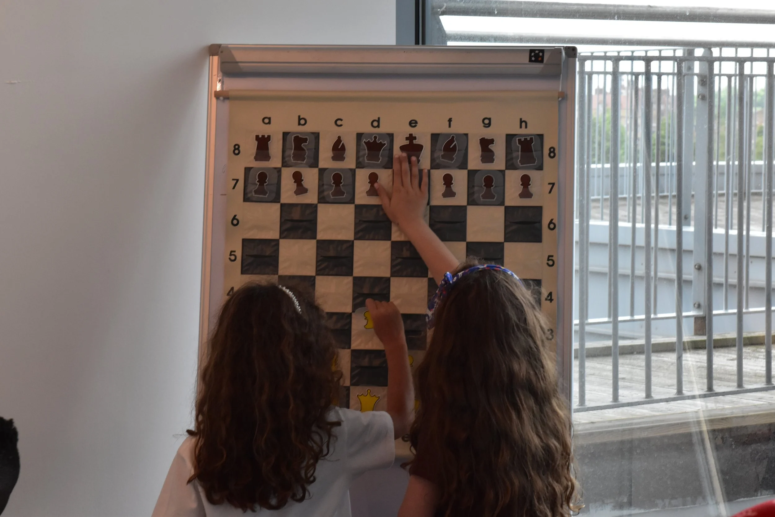 Children playing chess on a large chessboard with chess piece stickers, indoors near a window with bars.