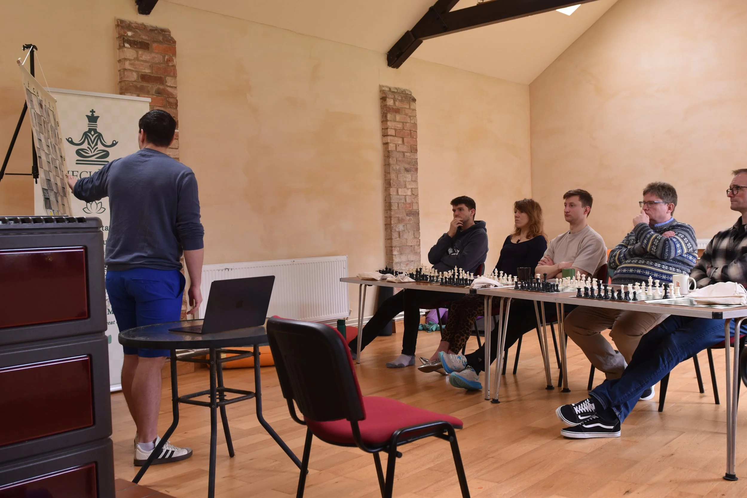 A group of six people sitting at a table playing chess, with one person standing near a display board in a room with beige walls and exposed brick columns.