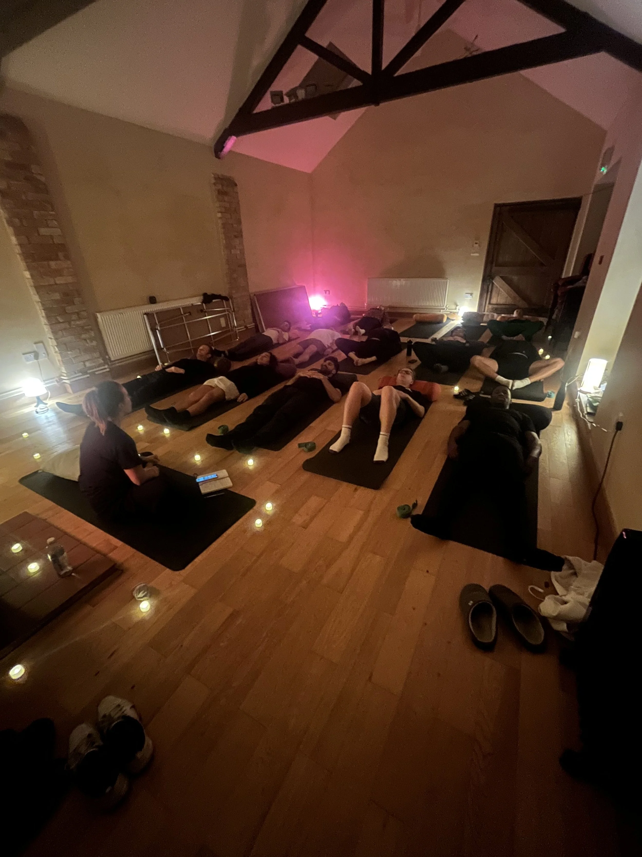 People participating in a yoga or meditation class lying on yoga mats in a dimly lit room decorated with candles, with a teacher at the front.