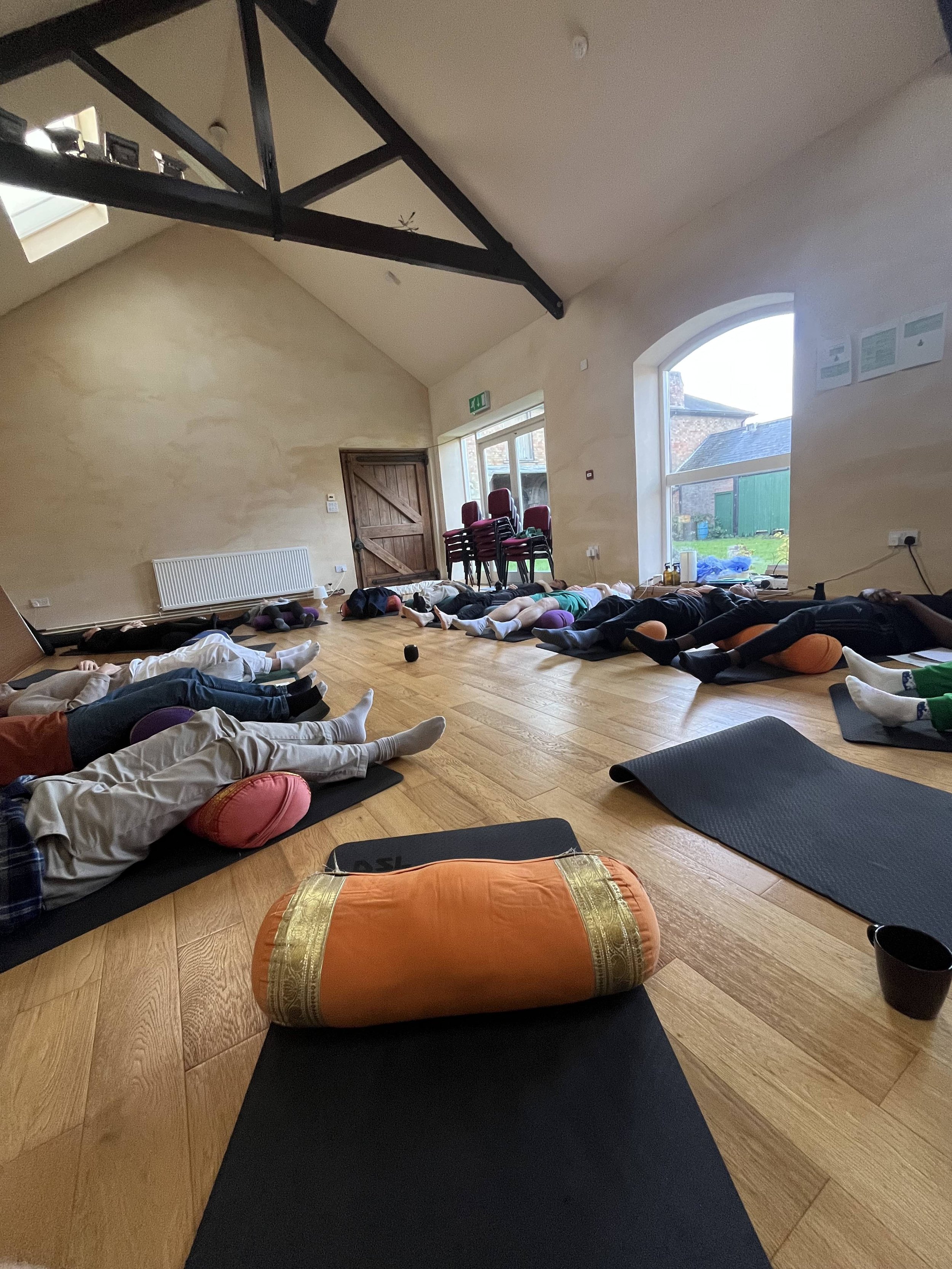 People lying on yoga mats in a meditation or yoga class inside a spacious room with wooden floors, large windows, and a sloped ceiling with black beams.