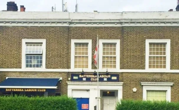 The exterior of the Battersea Labour Club, a two-story brick building with four windows on the upper floor and two on the lower, featuring a sign with the club's name and flags, surrounded by green bushes.