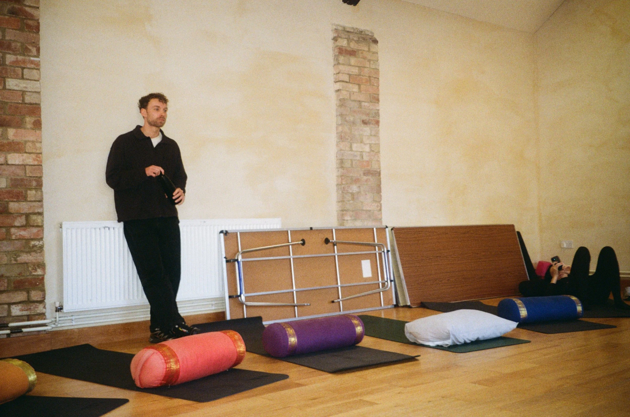A man standing in a yoga or exercise studio with mats and foam rollers on the floor, and a woman lying on her back using her phone in the background.