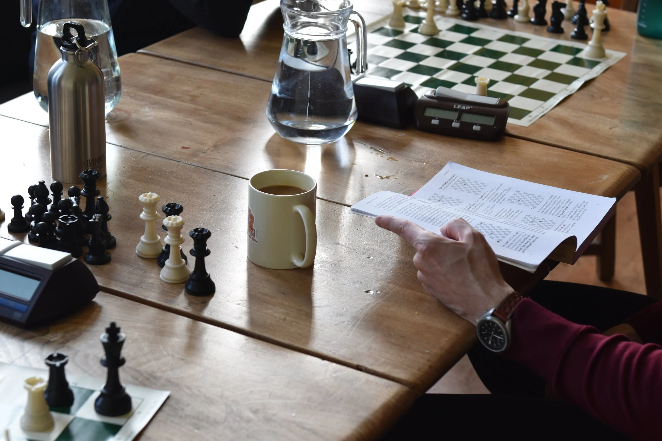 A wooden table set up for a chess game with a chessboard, chess pieces, a digital chess clock, a glass of water, a coffee mug, and a sheet of paper with chess instructions or analysis. There is a person's hand pointing at the sheet, and a wristwatch 