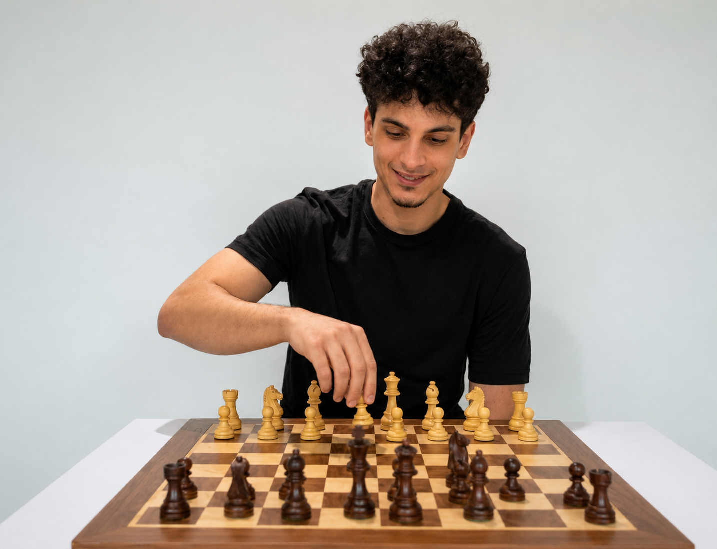 Young man playing chess on a green and white chessboard, smiling, wearing a black t-shirt, with a white background.