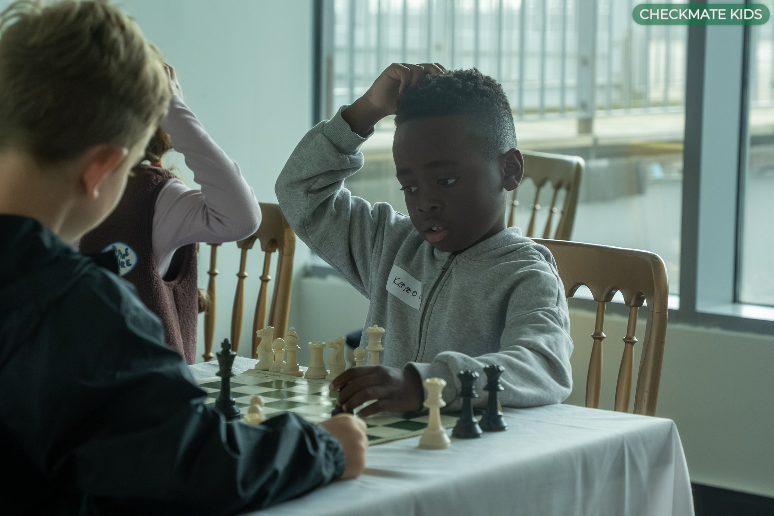 Children playing chess at a table indoors during daytime, with a young boy in a gray hoodie making a move and a girl with a button badge and a sweater observing, in a bright room with large windows.