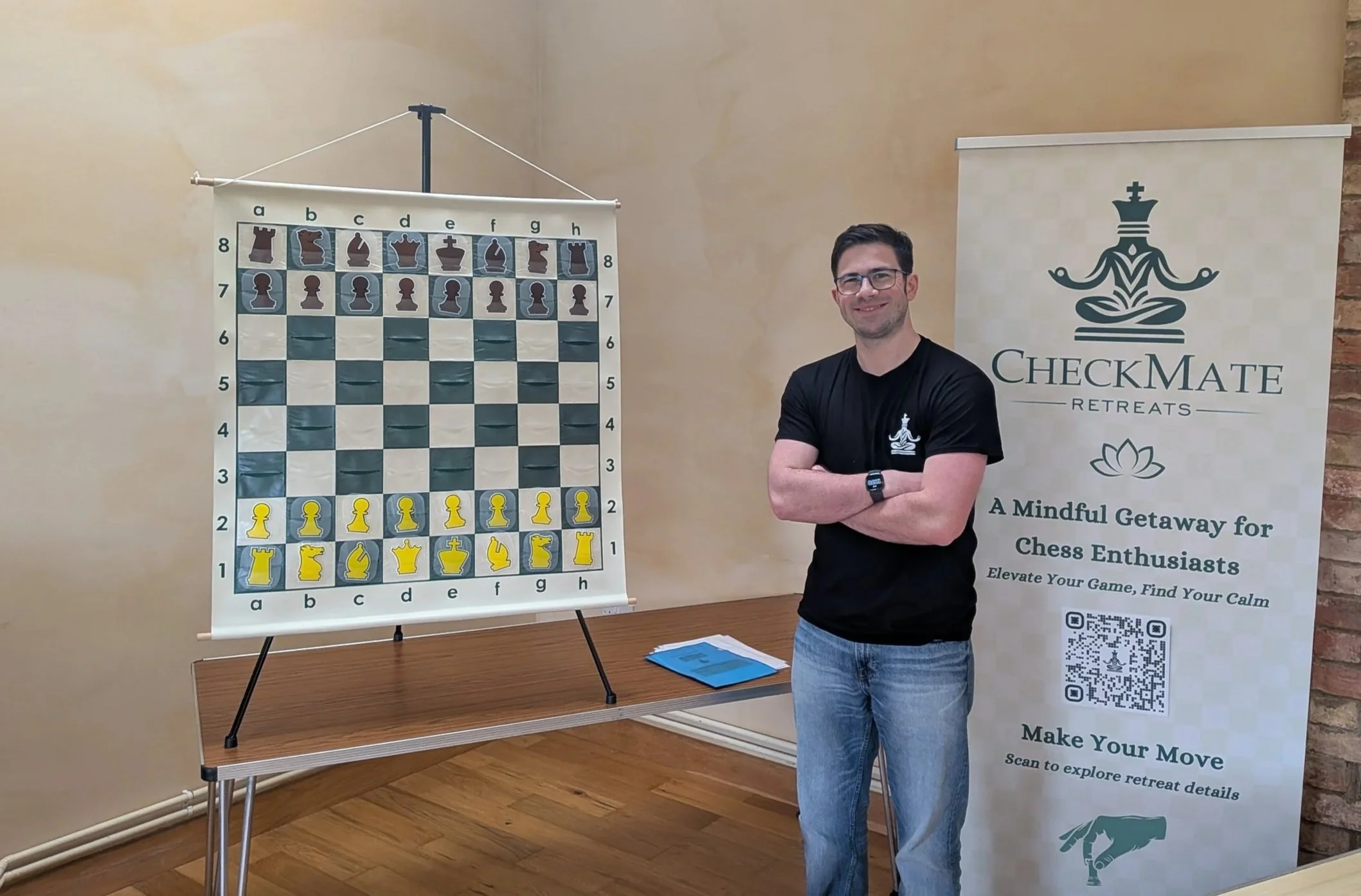 A man with glasses, wearing a black T-shirt and jeans, stands with arms crossed next to a large chessboard and a promotional banner for CheckMate Retreats. The chessboard has yellow and brown pieces set up for a game.