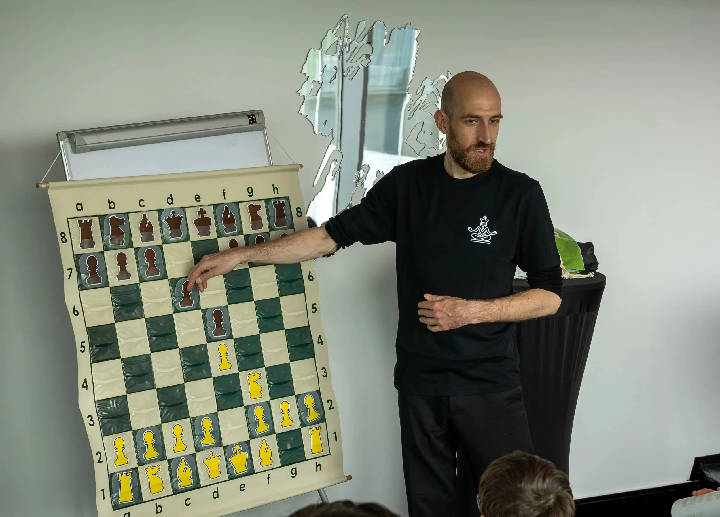 A man with a beard and shaved head in a black shirt standing next to a large green and yellow chessboard with chess pieces. The man is pointing to a location on the board, likely explaining a chess strategy. There is a mirror behind him reflecting part of the room, a whiteboard or flip chart, and some children seated in front, looking at the board.