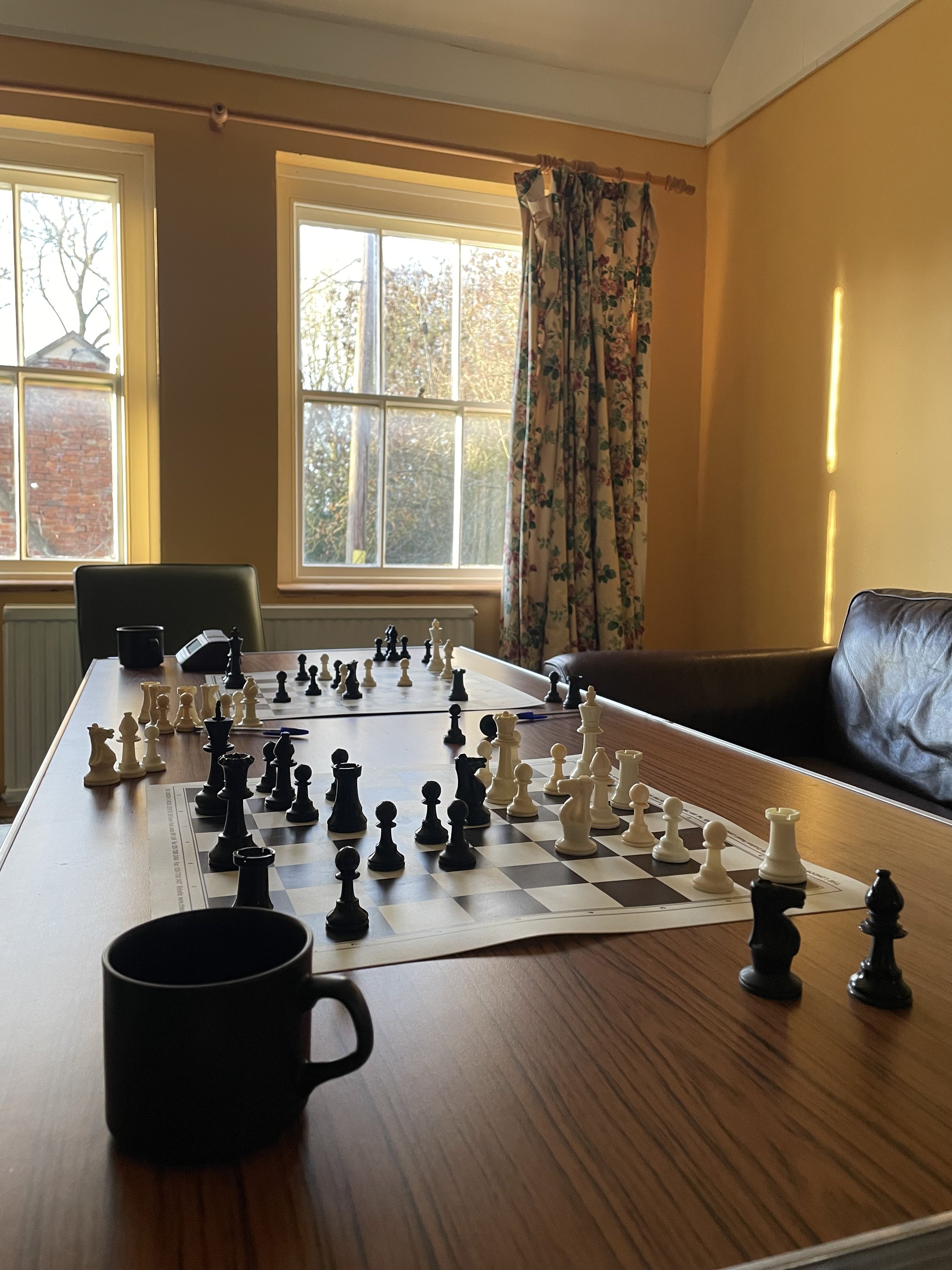 A chessboard set up for a game with black and white pieces on a wooden table. A black mug is in the foreground, and a window with sunlight shines through floral curtains in the background.