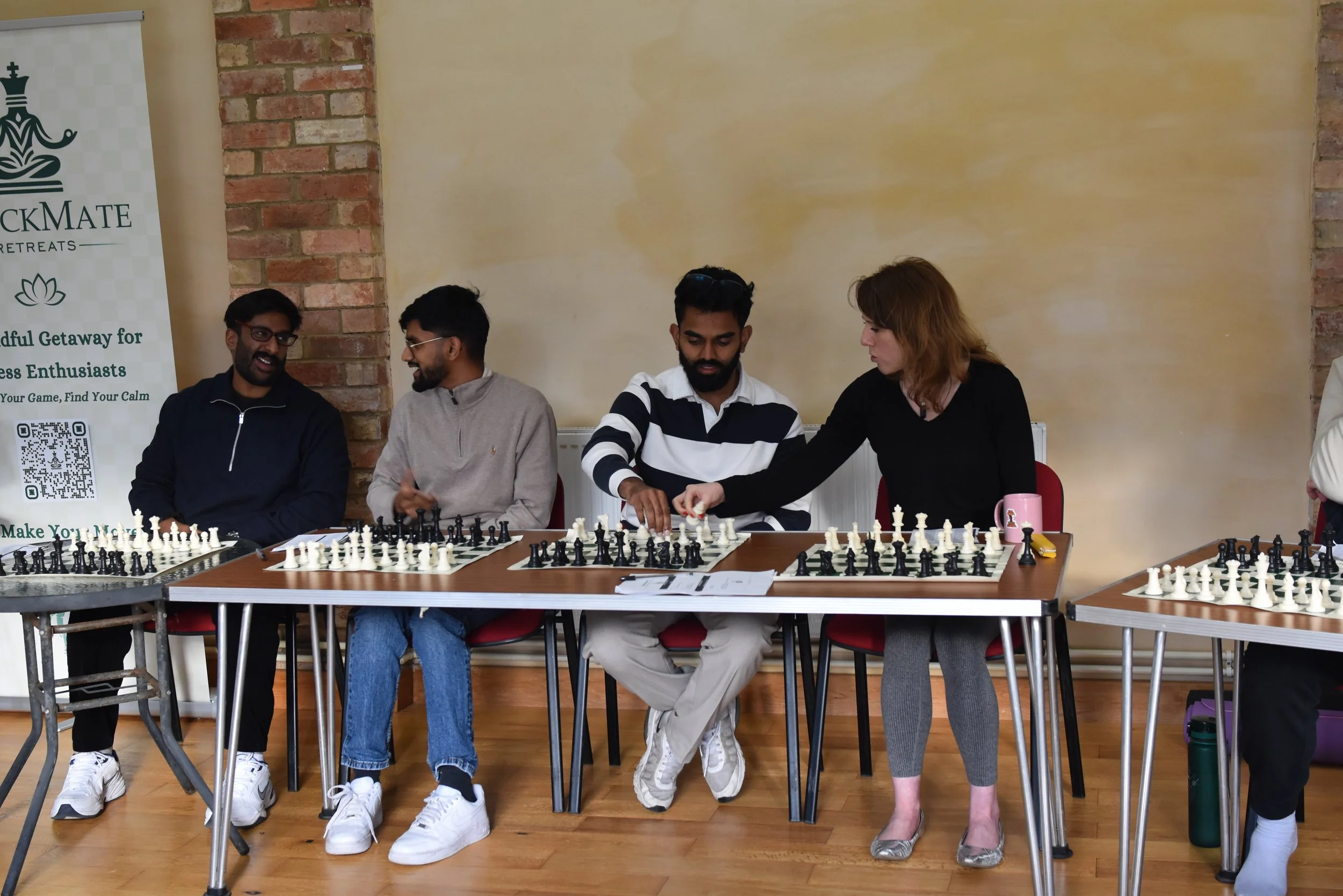 Four people playing chess at a table with a woman assisting. Behind them is a banner with text and a brick wall.