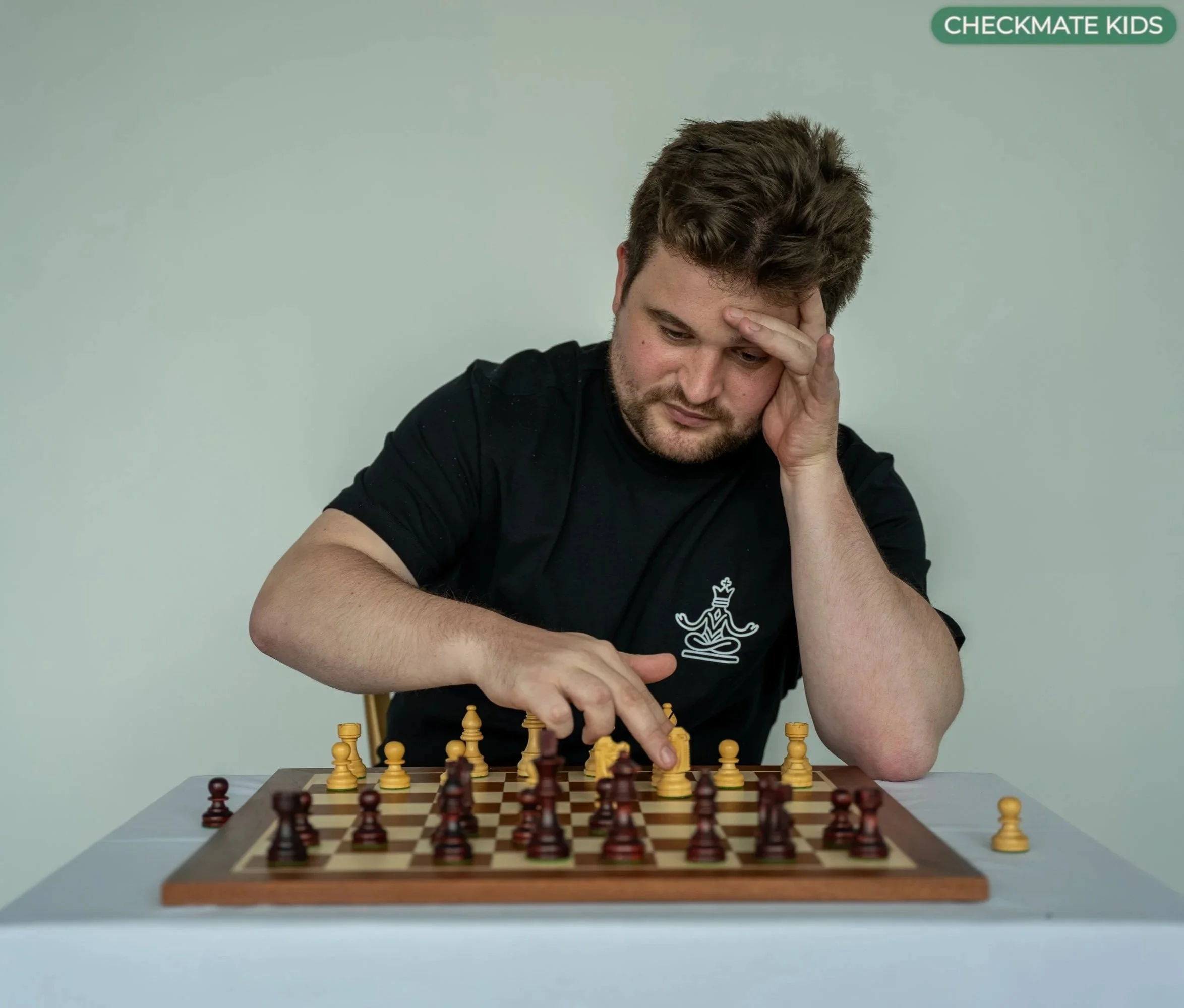 Young man playing chess, concentrating with hand on forehead, sitting at a chessboard.