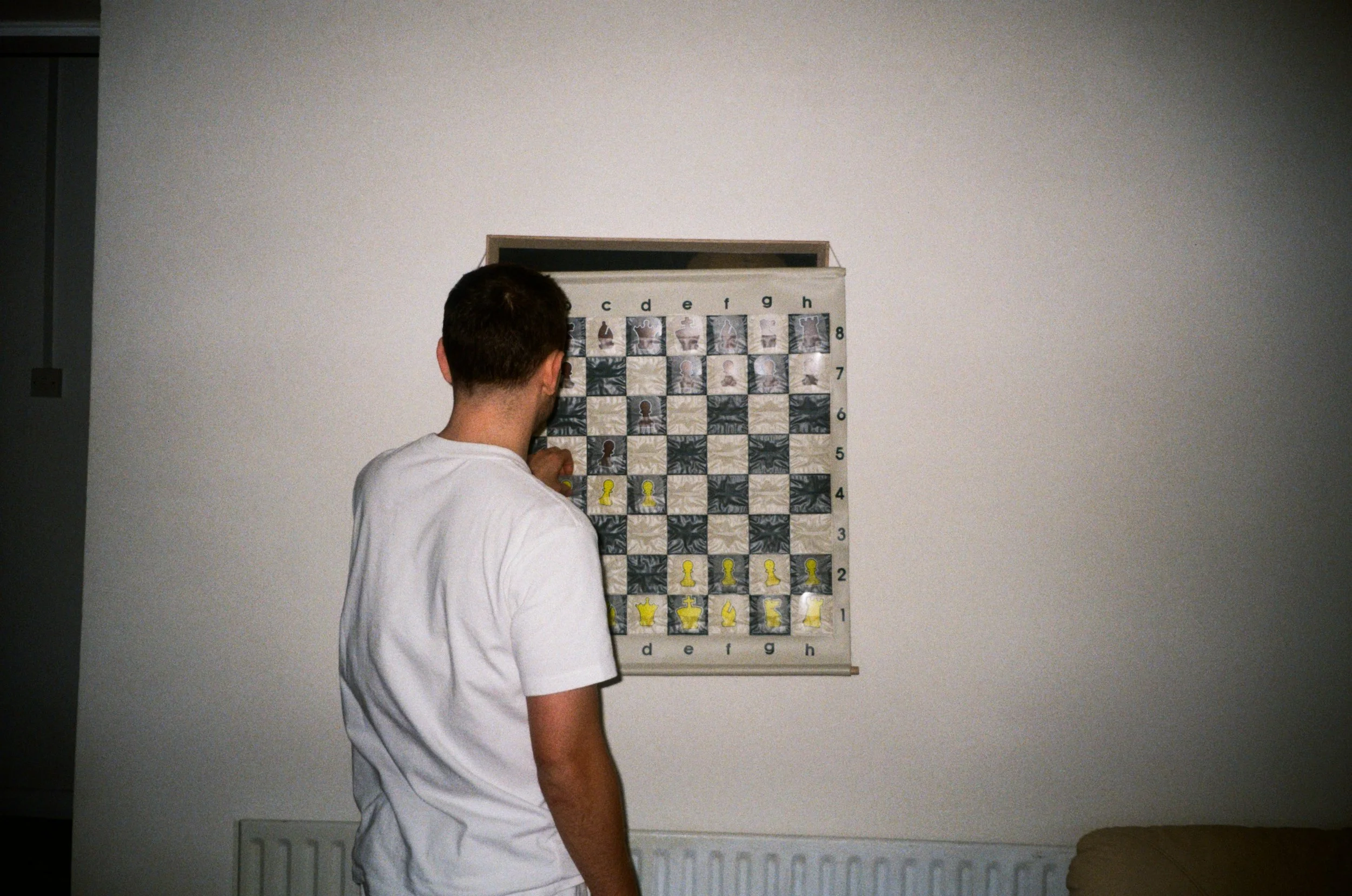 A young man in a white t-shirt playing chess on a wall-mounted chessboard.
