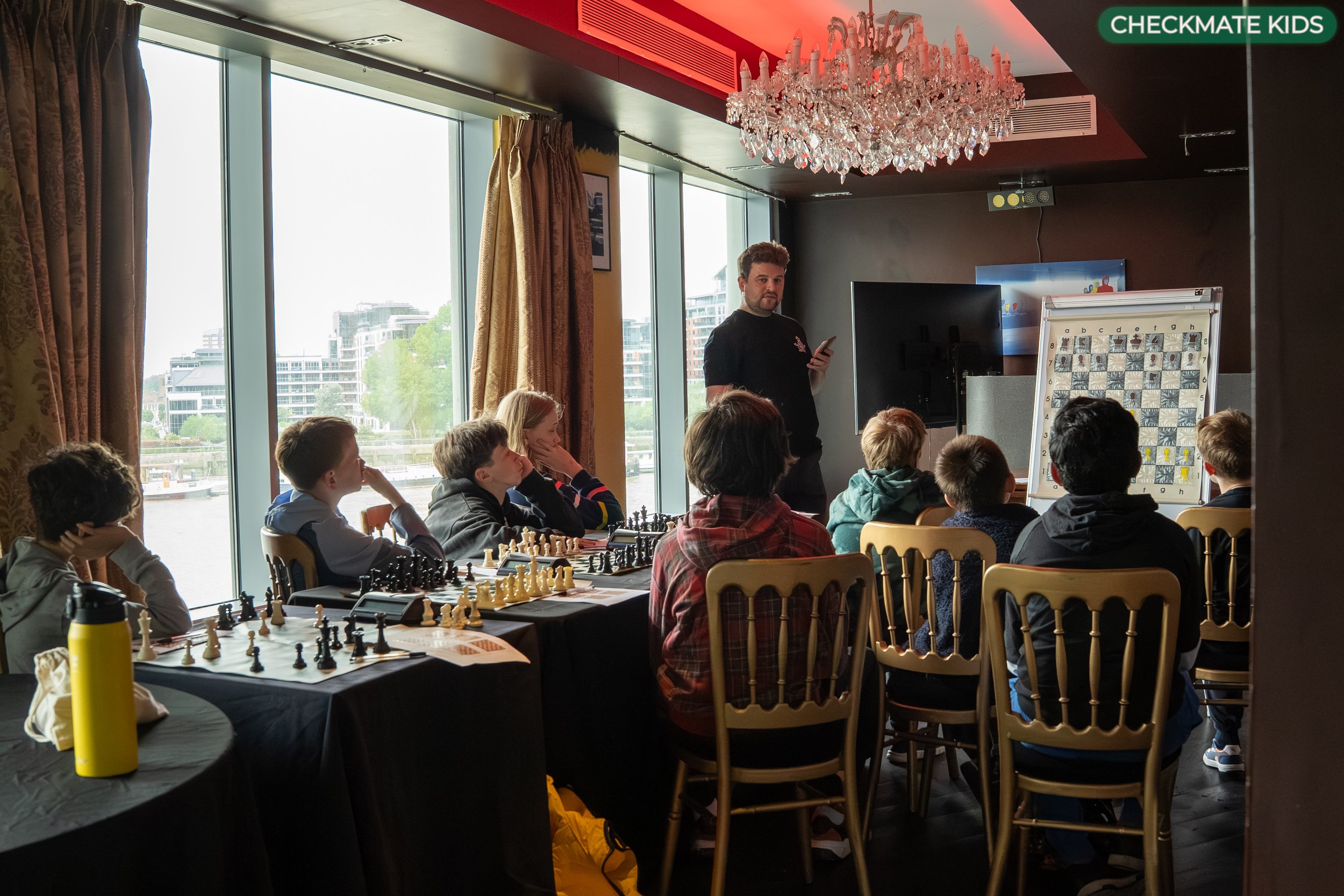 Group of children playing chess in a brightly lit room with large windows and city view, with an adult standing nearby, holding a phone, and a chandelier hanging from the ceiling.
