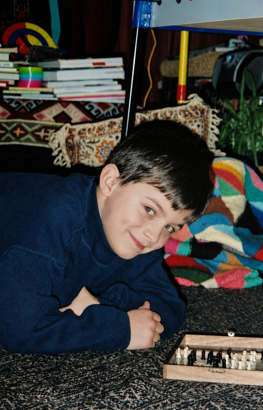 A young boy with dark hair and light eyes, lying on a patterned carpet, playing chess in a cozy room with a colorful blanket, books, and shelves in the background.