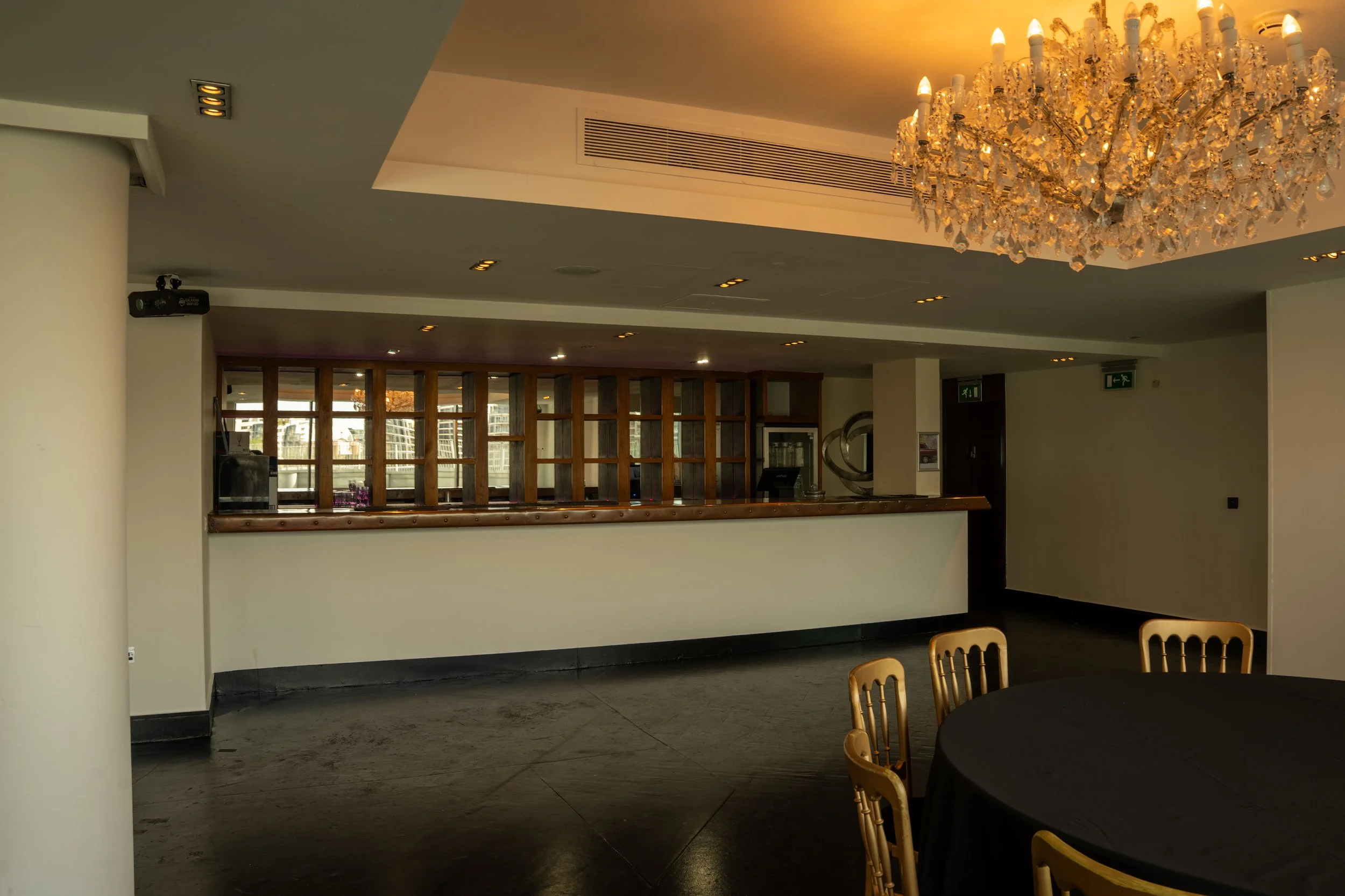 Interior view of a hotel lobby or event space with a large chandelier, a reception desk, and wooden furniture. The space has a modern design with a high ceiling and windows in the background.