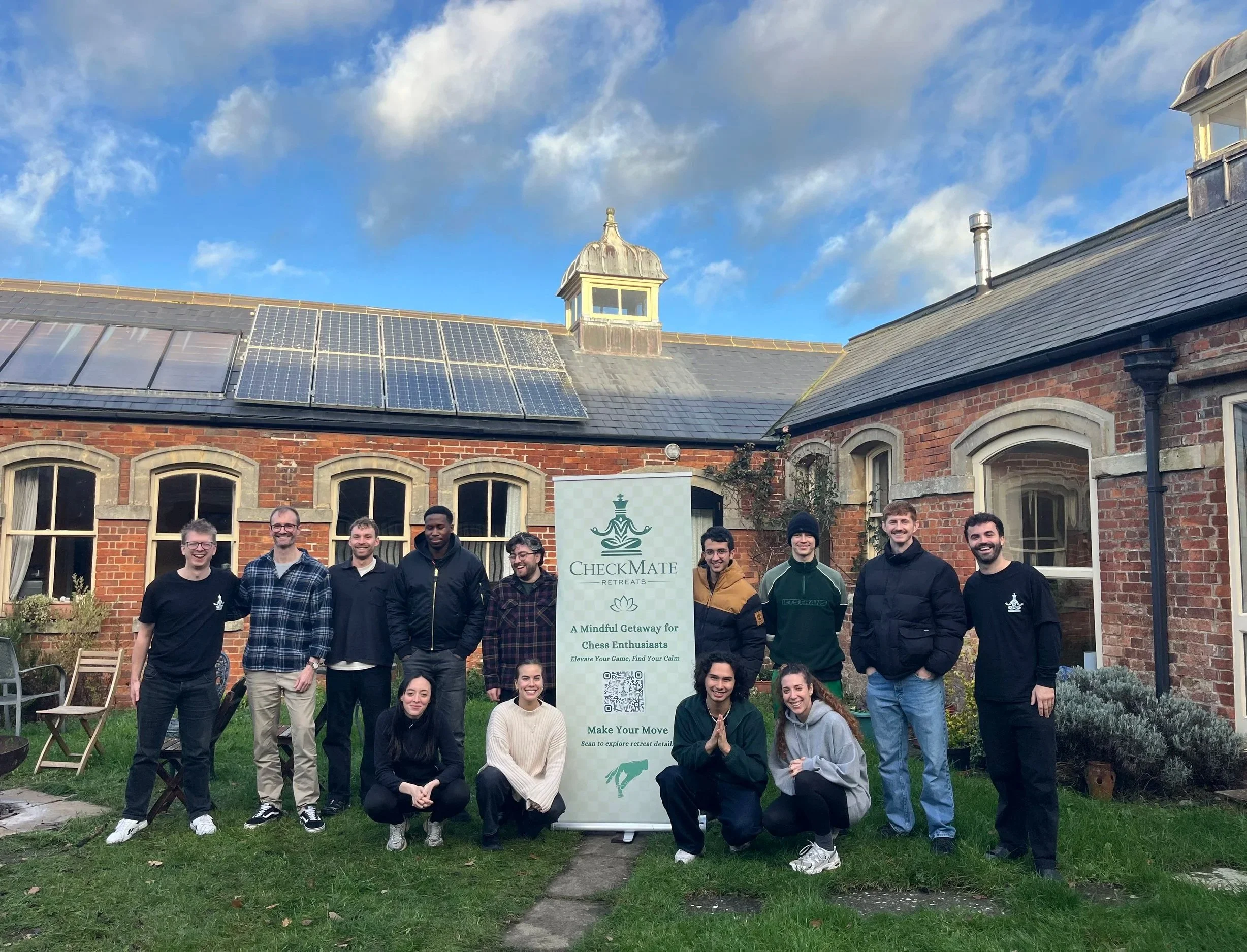 A group of people standing and sitting outdoors in front of a brick building with roofing solar panels and a sign that reads 'Checkmate Retreats'. The sky is partly cloudy.