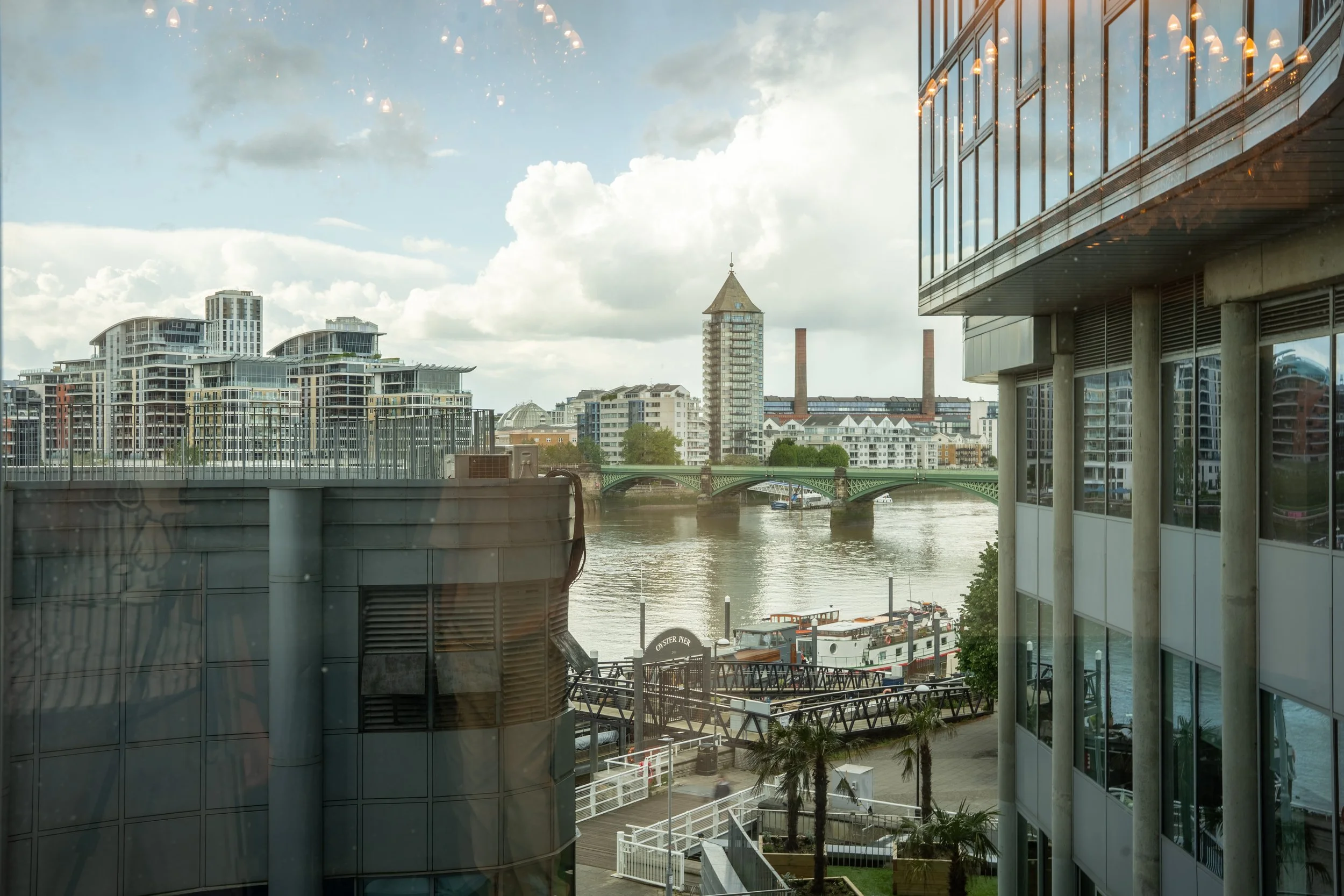 Cityscape view through glass windows, featuring modern buildings, an old tower, a bridge over a river, and boats docked near the waterfront.